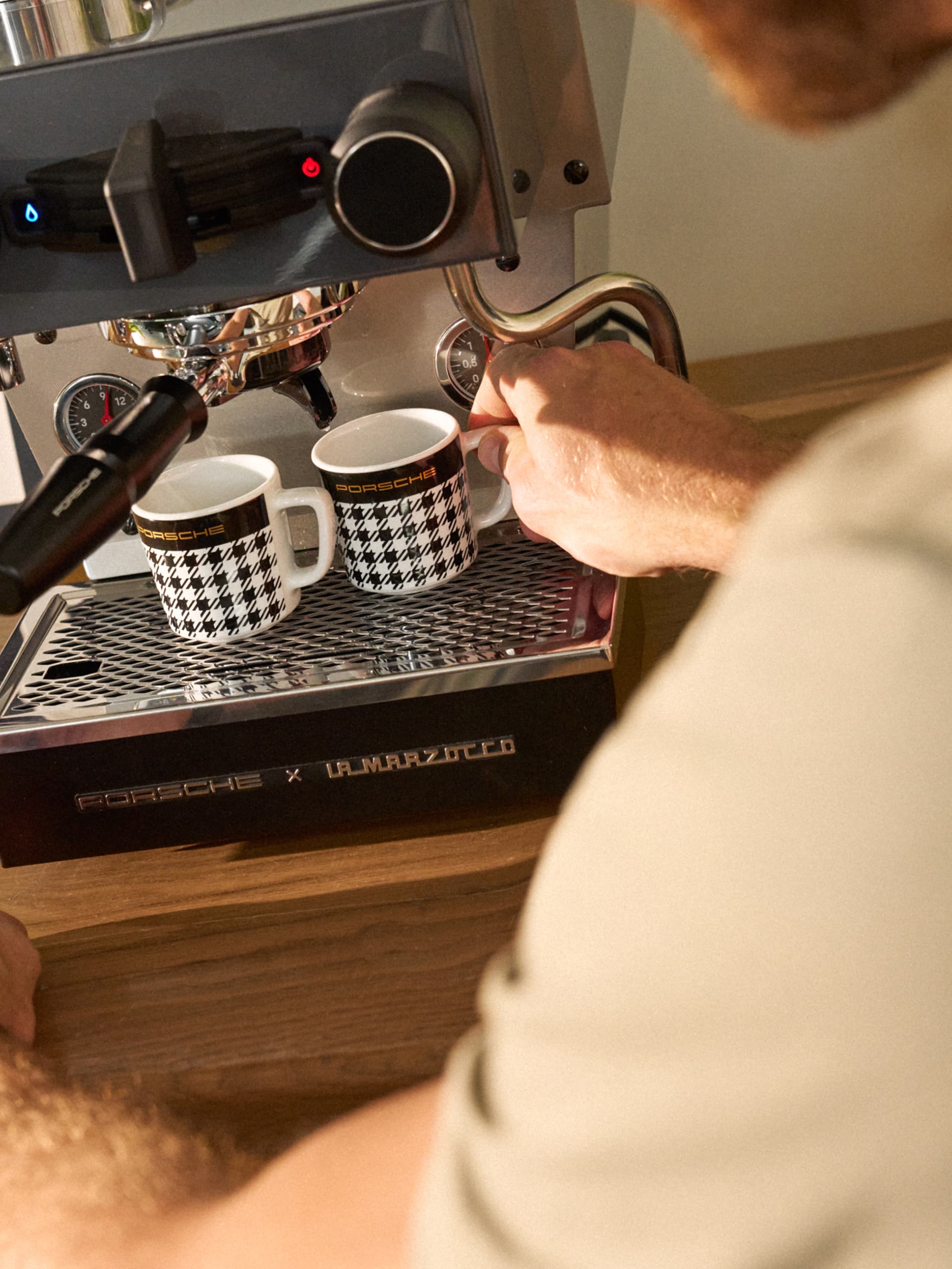 Main plaçant une tasse à espresso Porsche à motif pied-de-poule sous une machine à café, avec une tasse assortie sur le plateau d’égouttage en métal.
