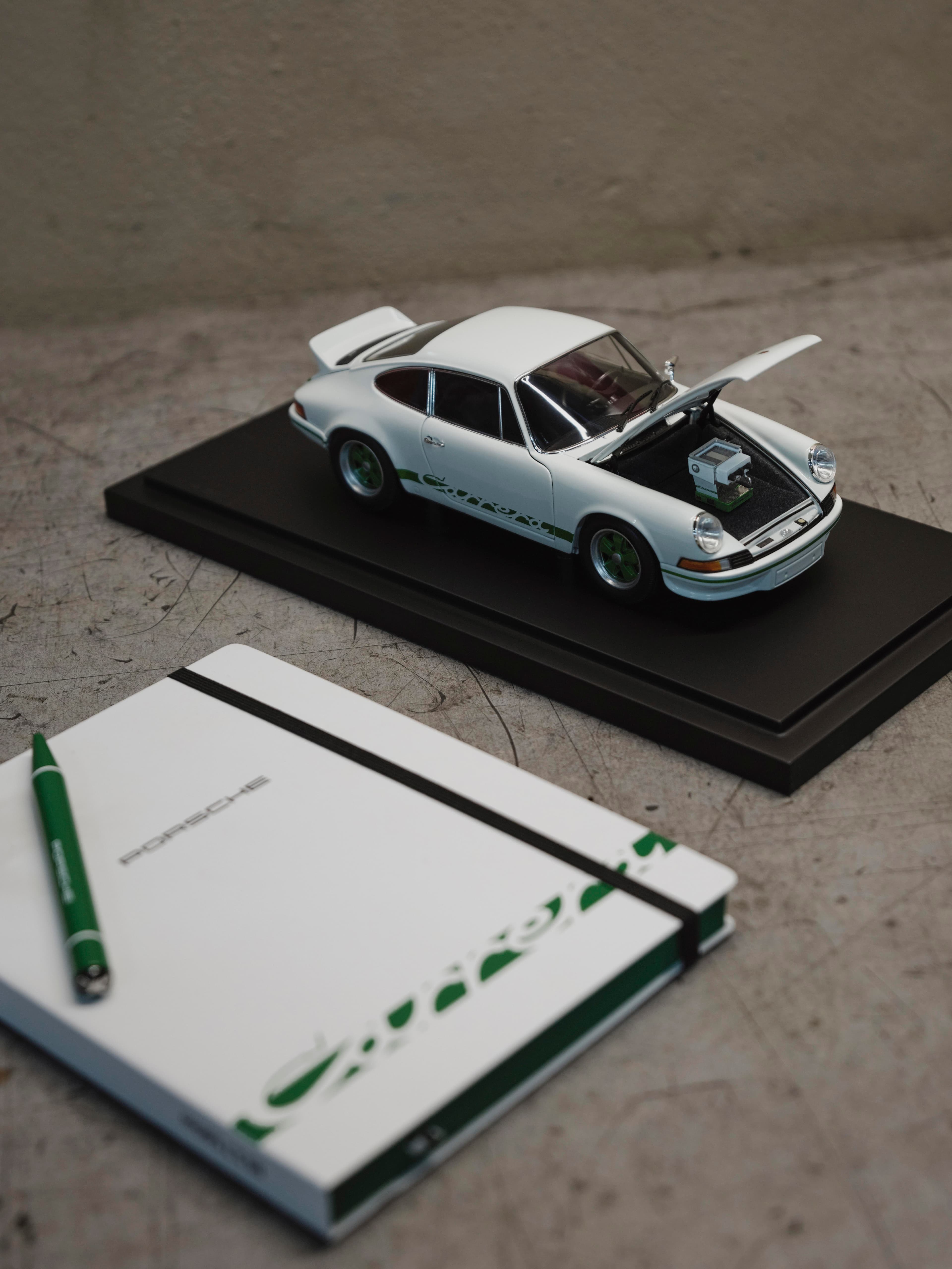 Weißes Porsche 911 Modellauto mit grünen Details auf einem Sockel, Motorhaube geöffnet; daneben Notizbuch und Stift.