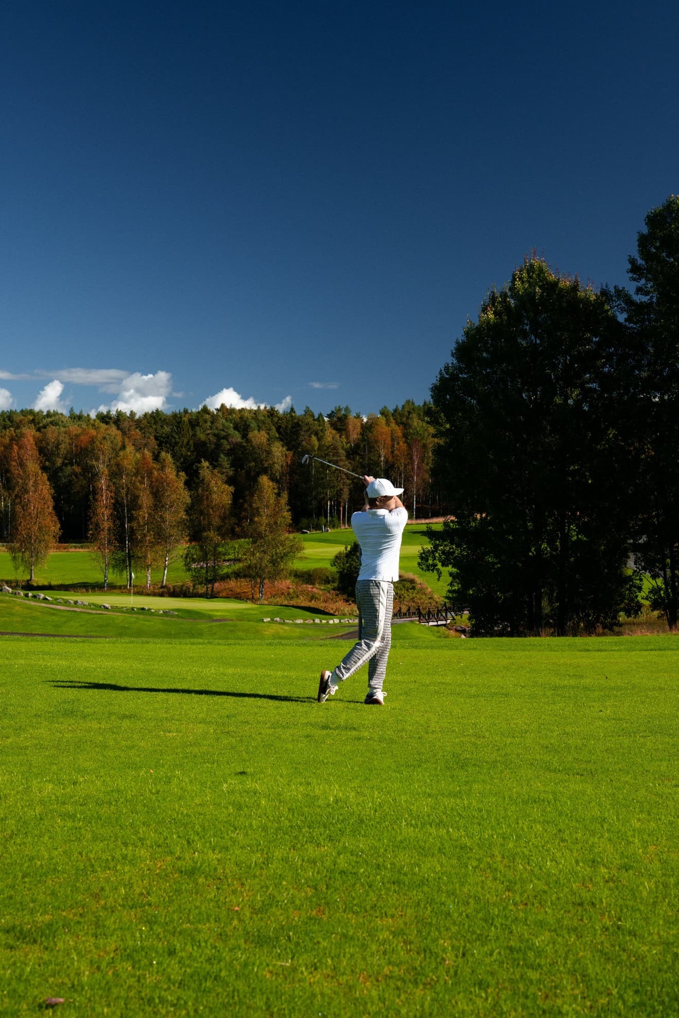 Golfista con cappellino e maglia bianchi mentre esegue uno swing su un fairway verde, alberi autunnali e cielo blu intenso sullo sfondo