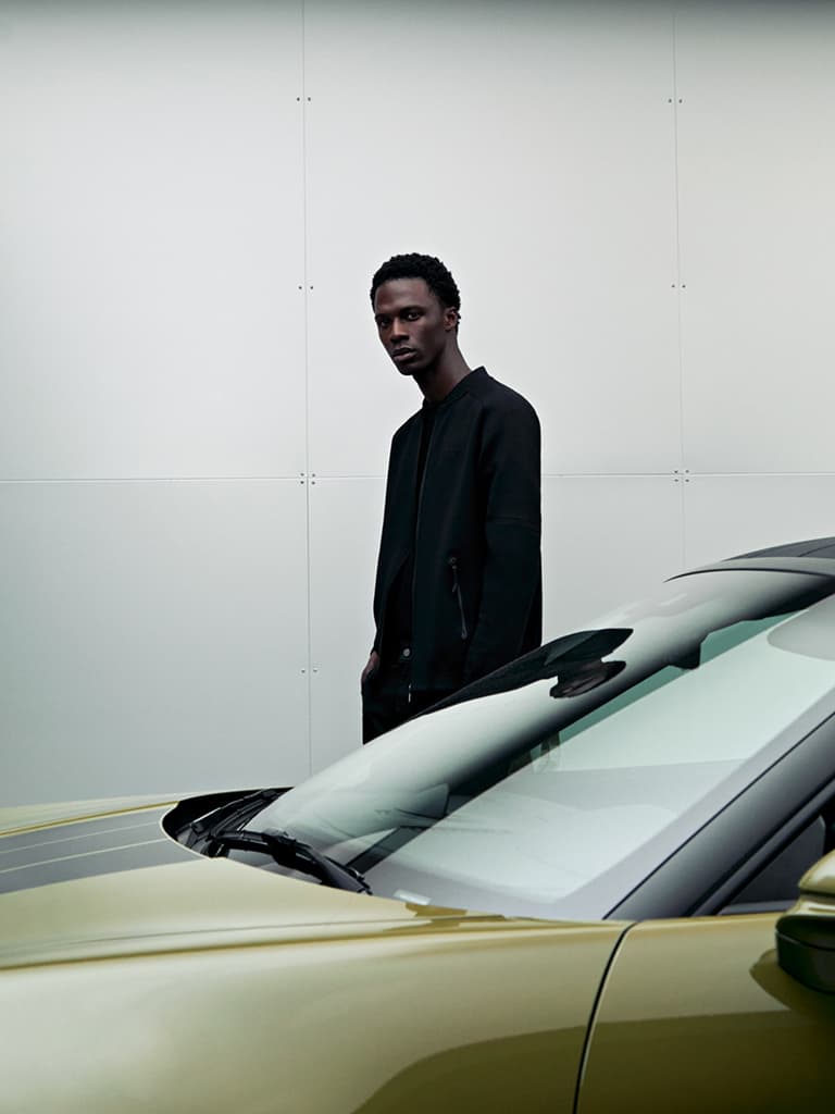 Homem com casaco preto em frente a uma parede branca; em primeiro plano, o para-brisas e o capô de um Porsche dourado.