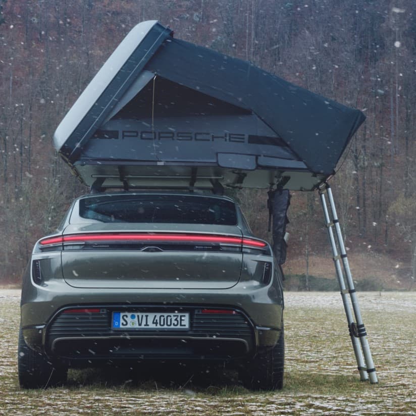 Porsche car with rooftop tent in snowy field, rear view, forest background, ladder attached to tent.