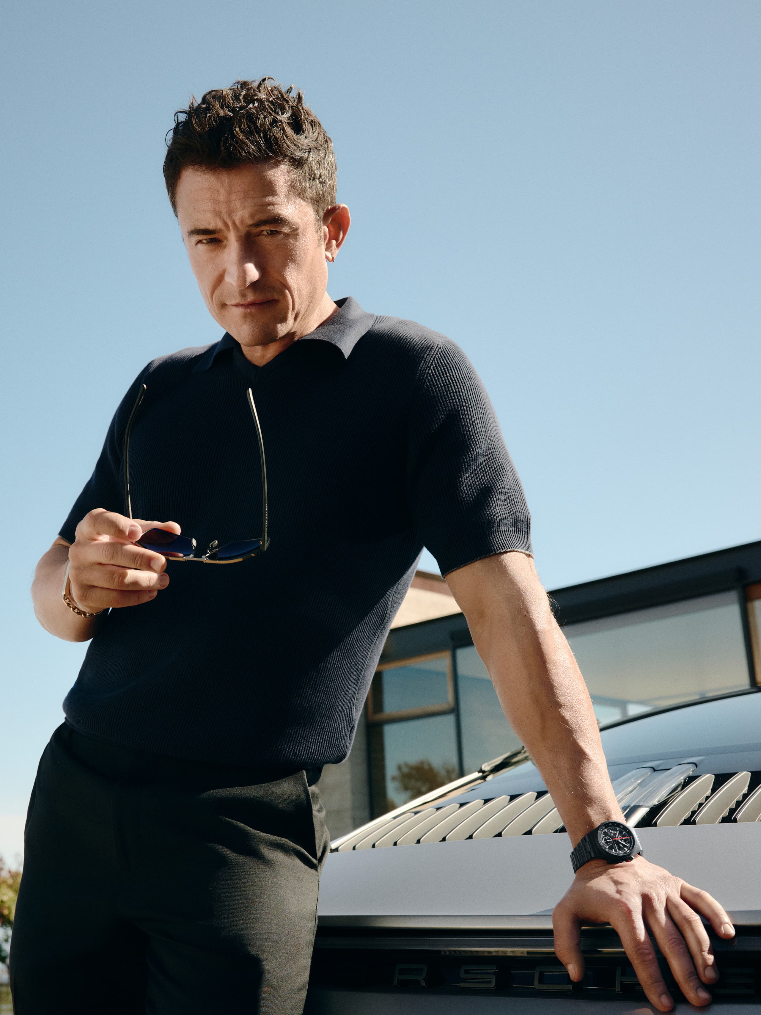 Orlando Bloom wearing the Chronograph 1 - All Black Numbered Edition on his wrist leaning against the rear of a Porsche.