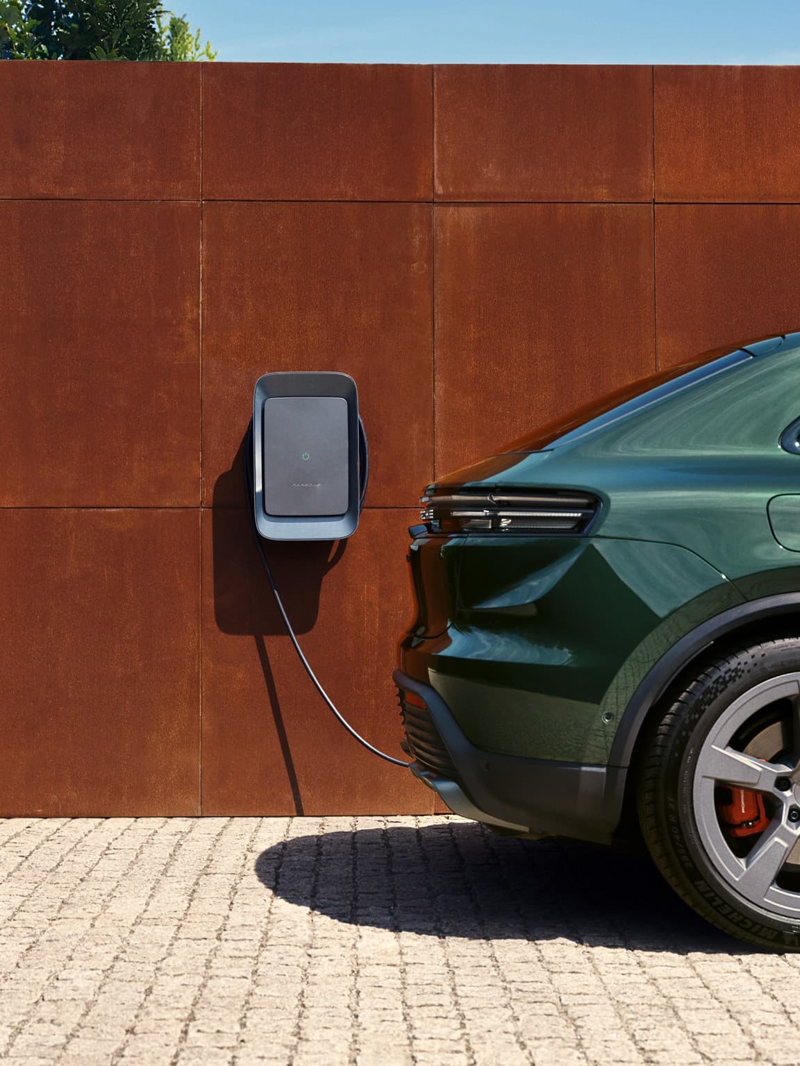 Green Porsche charging at a wall-mounted station on a rust-colored wall, sunny day, cobblestone ground.