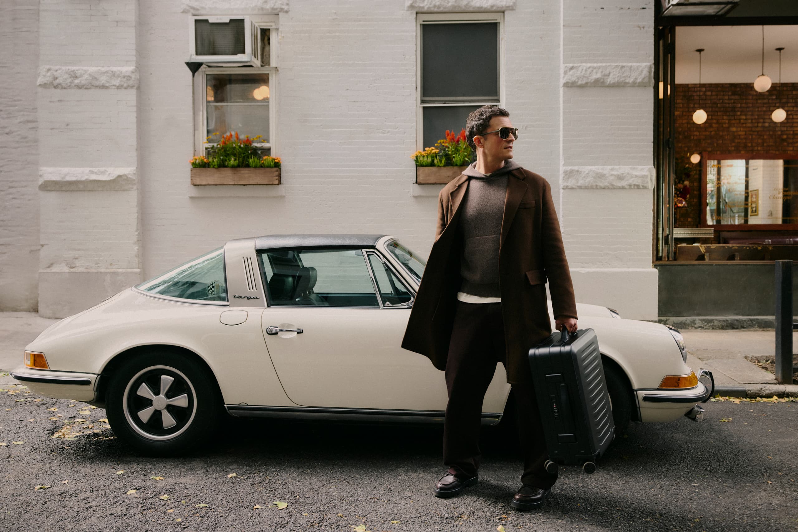 Orlando Bloom z aluminiową walizką na kółkach Porsche Design Roadster, w tle samochód Porsche