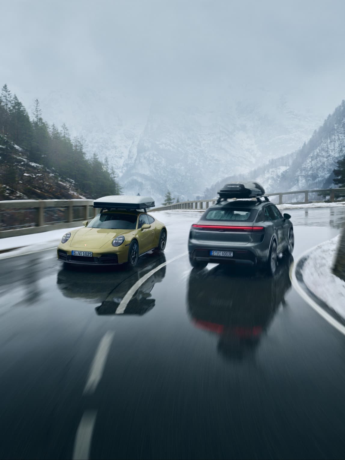 Two Porsche cars, yellow and gray, drive on a wet mountain road with snow-covered trees and misty mountains in the background.