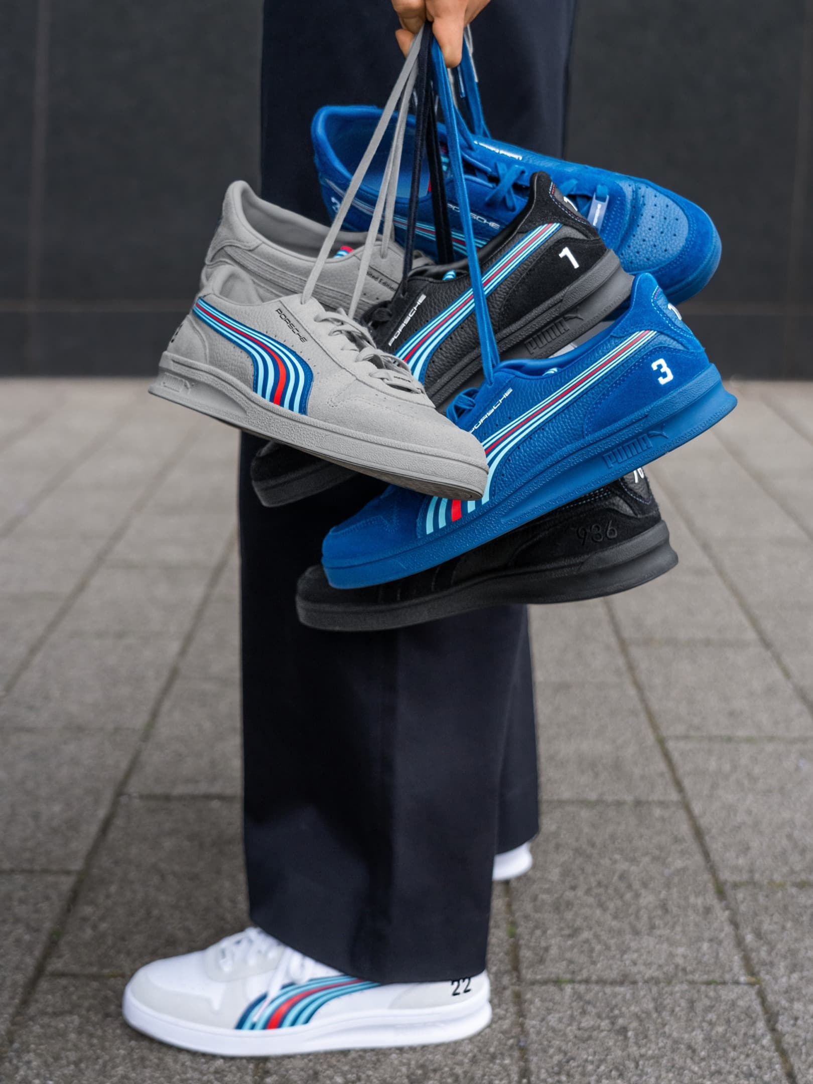 Person holding gray, black, and blue Porsche sneakers with striped design; wearing white sneakers on a tiled floor.