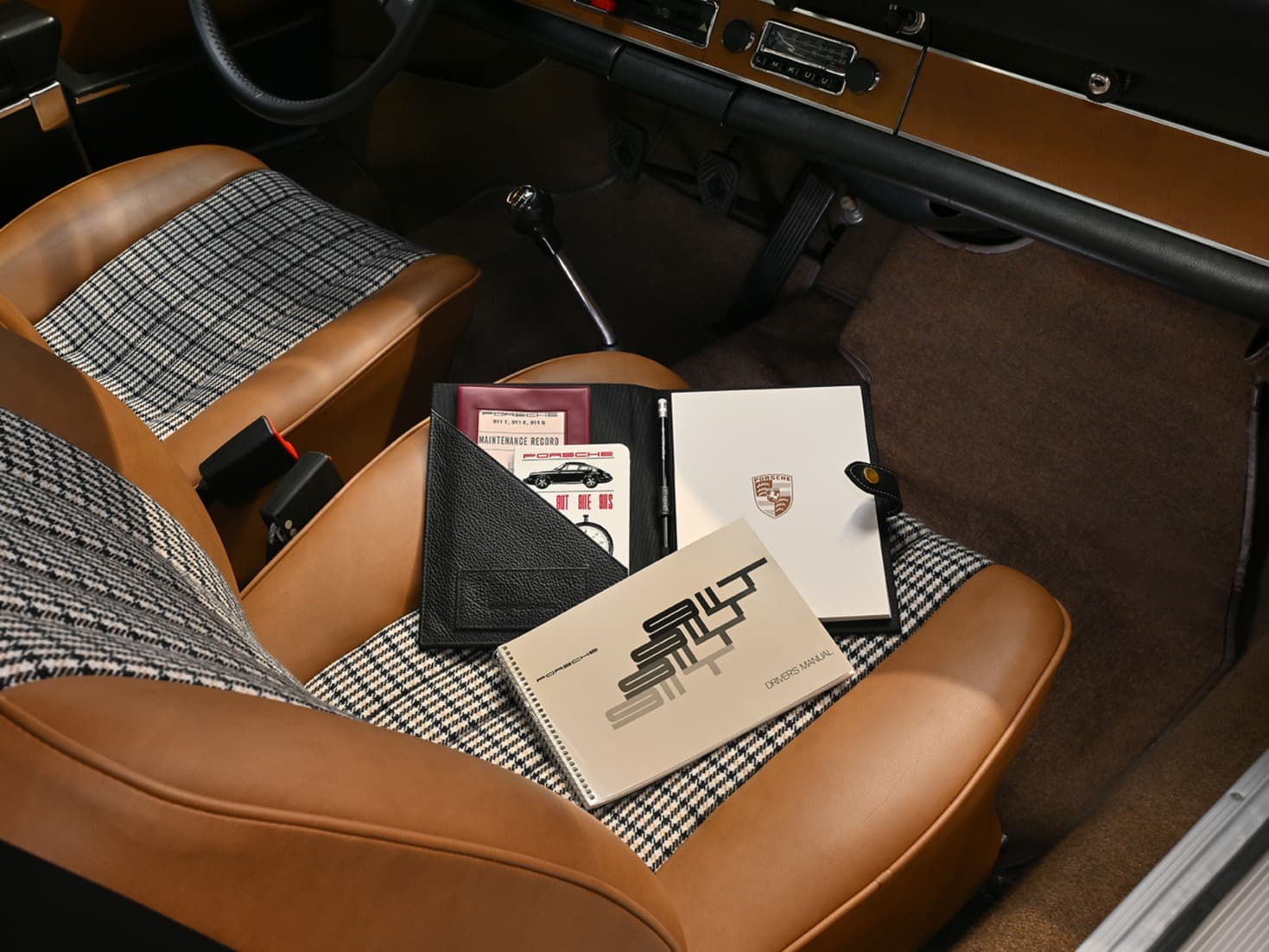 Porsche-bilinteriør med lysebrunt skinn og houndstooth-seter, manualer og dokumentmappe på passasjersetet