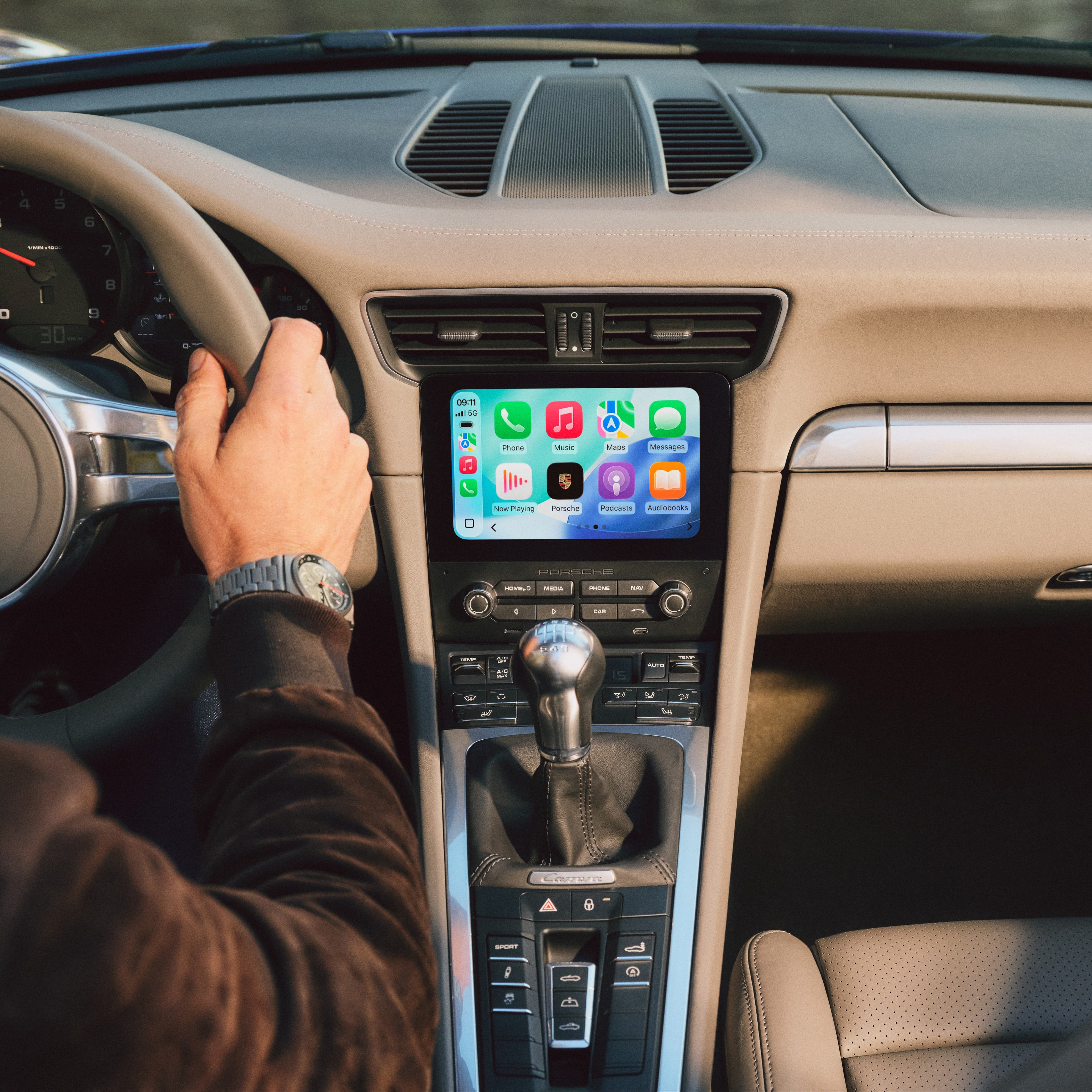 Förare i beige Porsche-interiör med händerna på ratten, mittmonterad pekskärm som visar Apple CarPlay-appar.