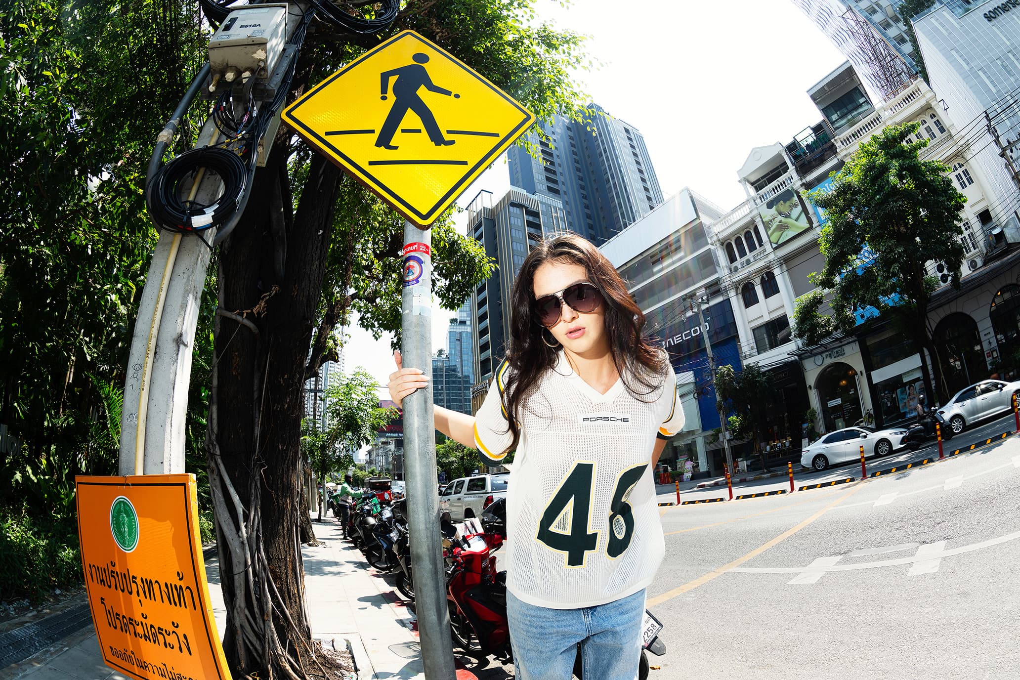 Woman wearing sunglasses in a white Porsche AHEAD football shirt by a yellow pedestrian crossing sign, city street in the background.