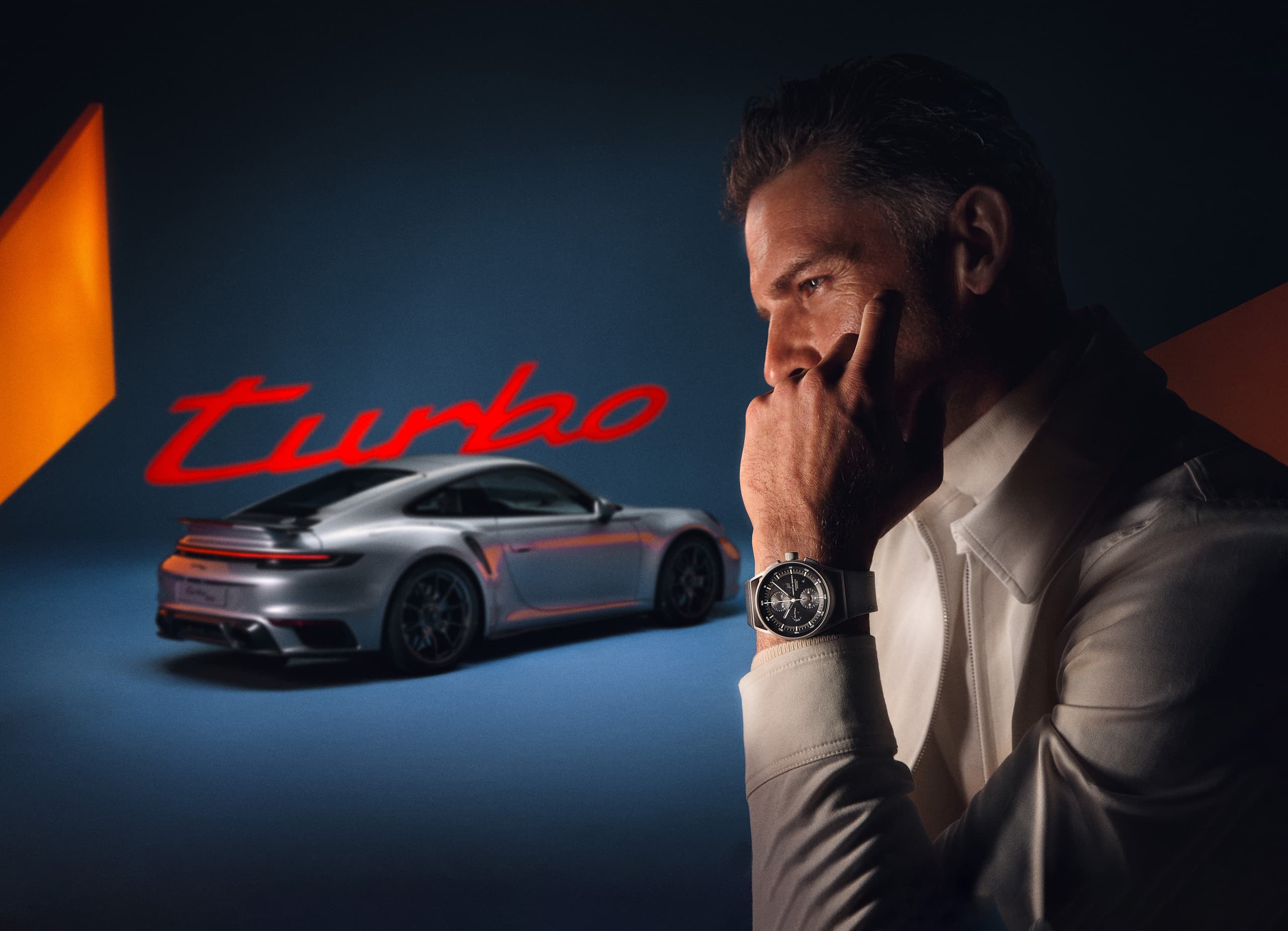 Mężczyzna z czarnym zegarkiem na rękę, widziany z profilu, w tle srebrne Porsche 911 Turbo i czerwony napis „turbo”.