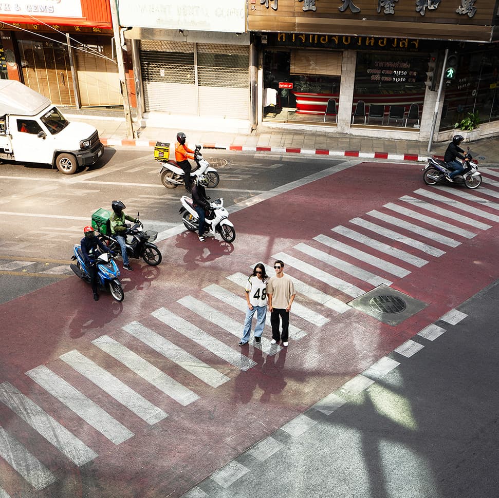 Two models wearing the Porsche AHEAD collection on a red zebra crossing, surrounded by motorcycles and a white delivery van in the city.
