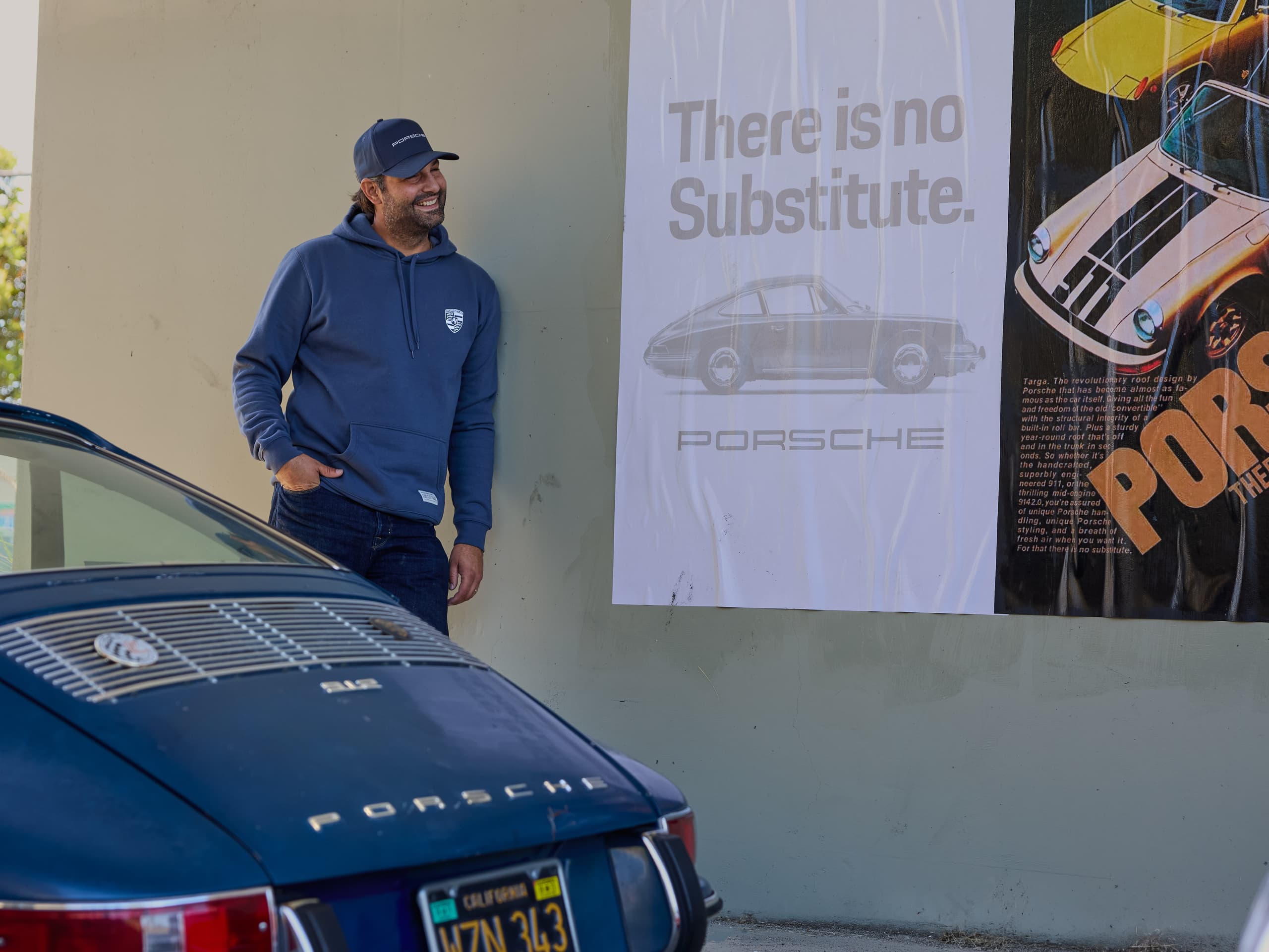 Man in a blue Porsche hoodie and cap leaning against a wall next to a Porsche poster; in the foreground, the rear of a blue Porsche.