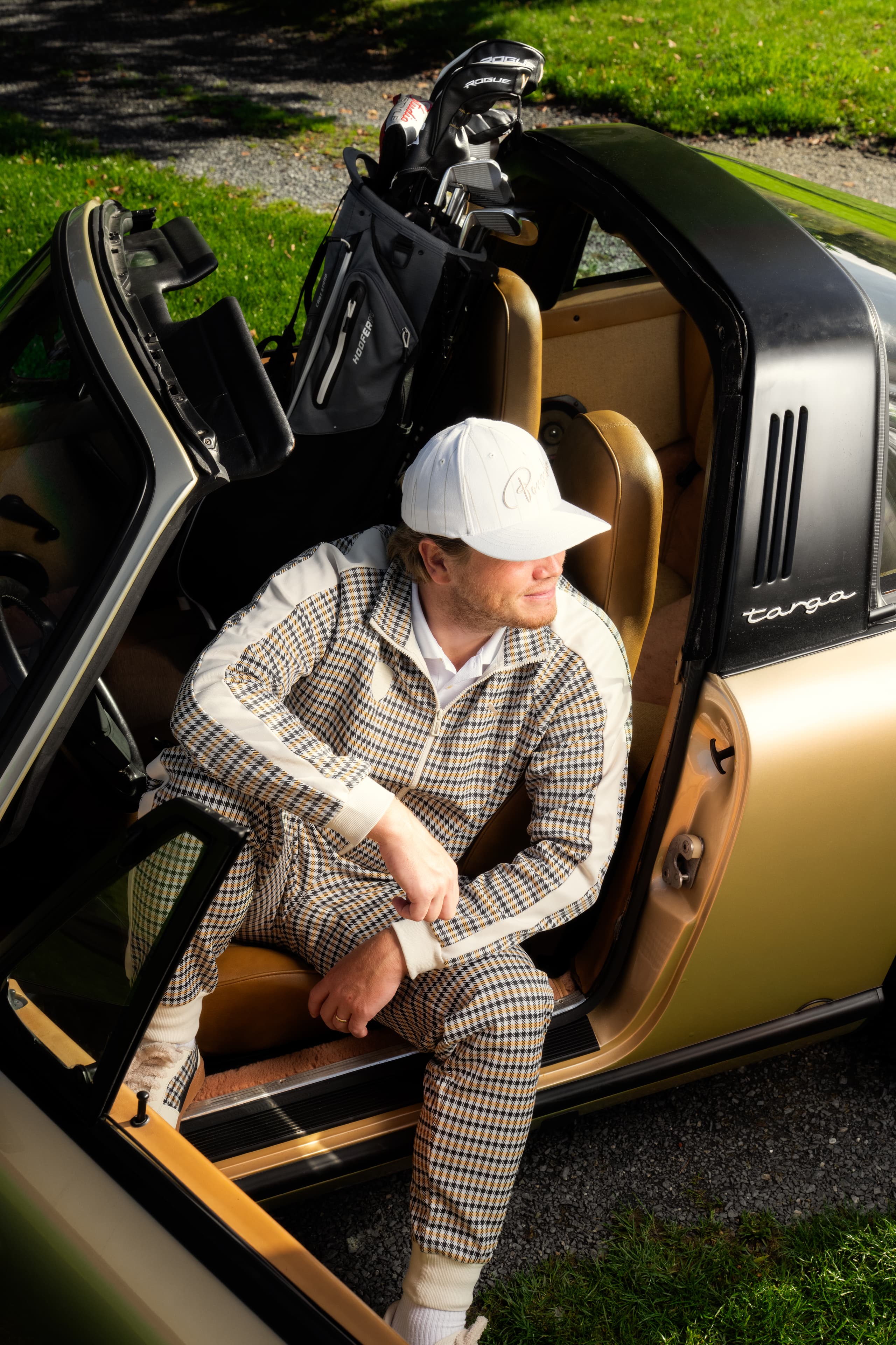 Hombre con chándal estampado y gorra blanca sentado en un Porsche Targa dorado descapotable, con una bolsa de golf detrás del asiento.