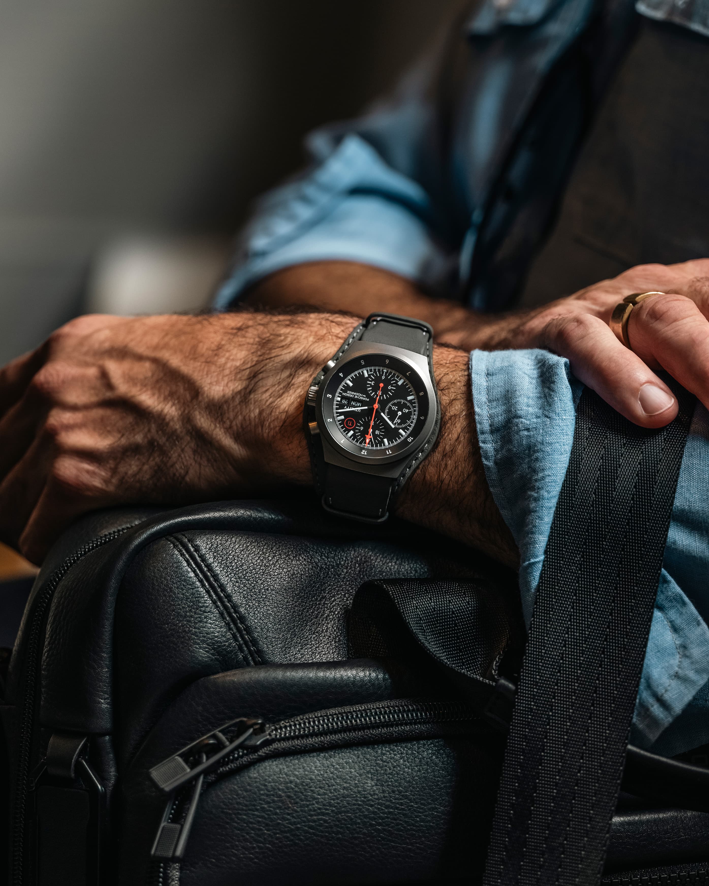 Primo piano di un polso che indossa un orologio cronografo nero con lancetta dei secondi rossa, appoggiato sulla tracolla di una borsa in pelle nera.