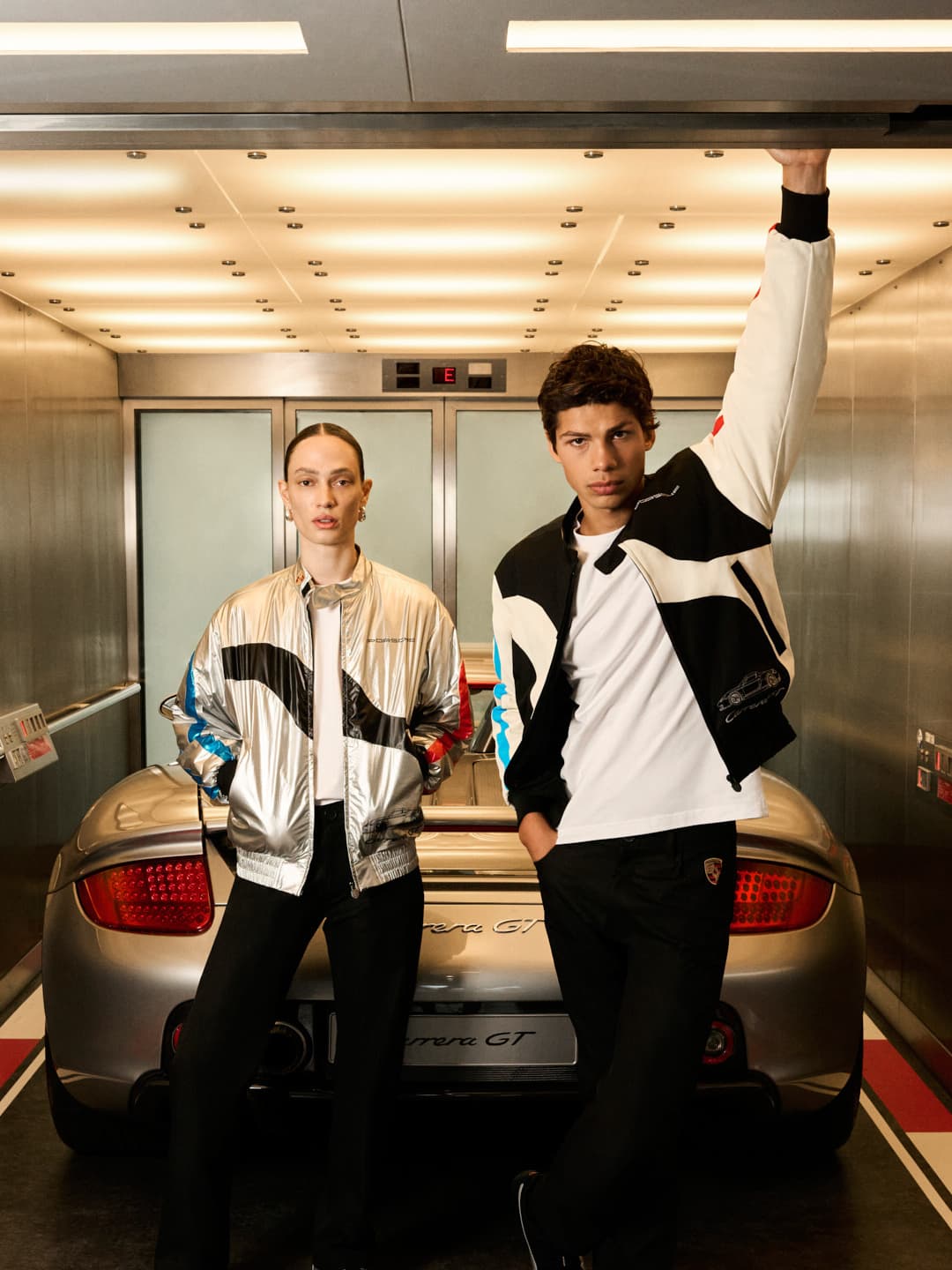 Woman and man wearing clothing from the Porsche Carrera GT 25th Anniversary Collection, standing in front of a Porsche Carrera GT