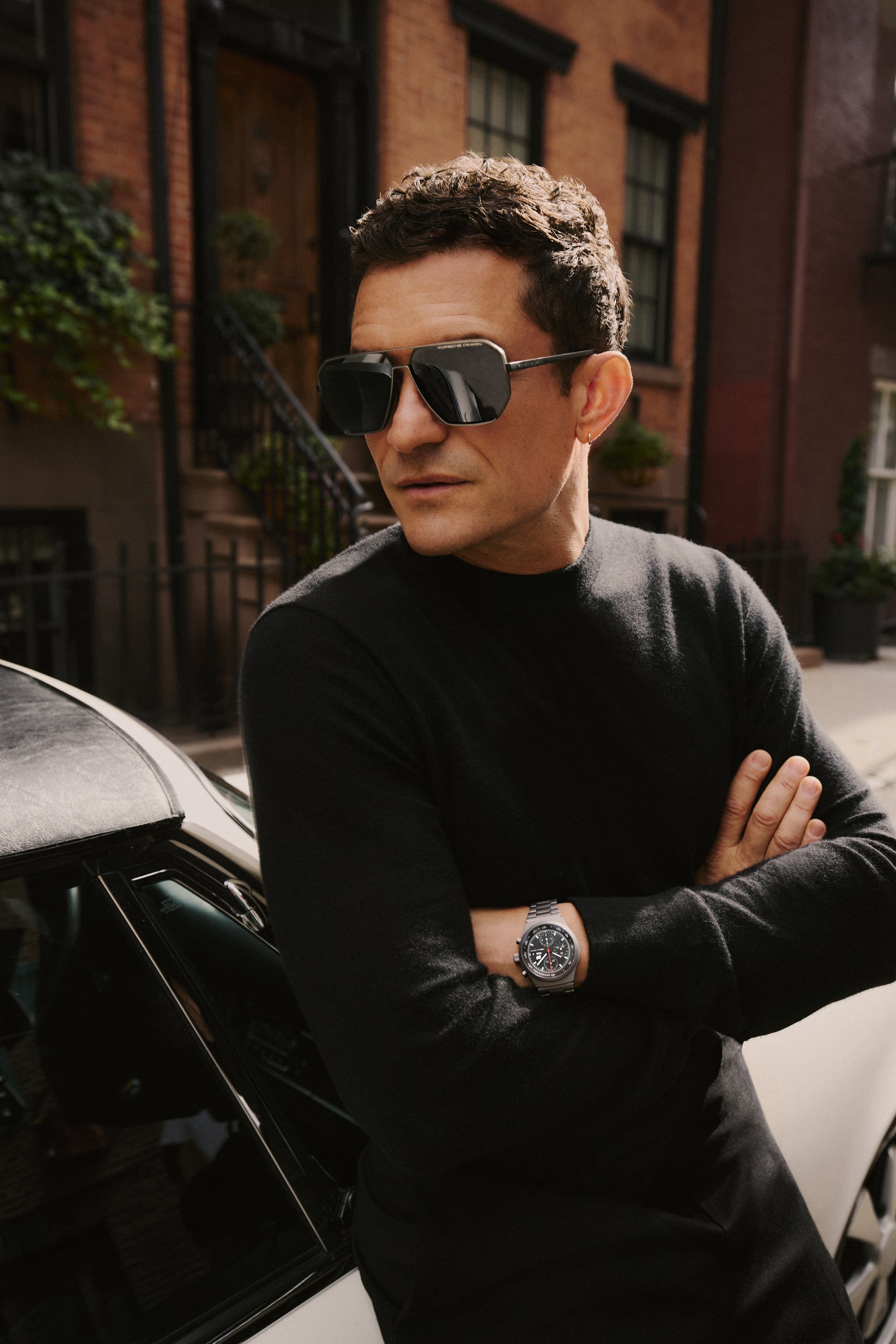 Orlando Bloom wears the Porsche Design Targa Eyewear series and stands with arms crossed on a sunlit residential street lined with brick buildings.