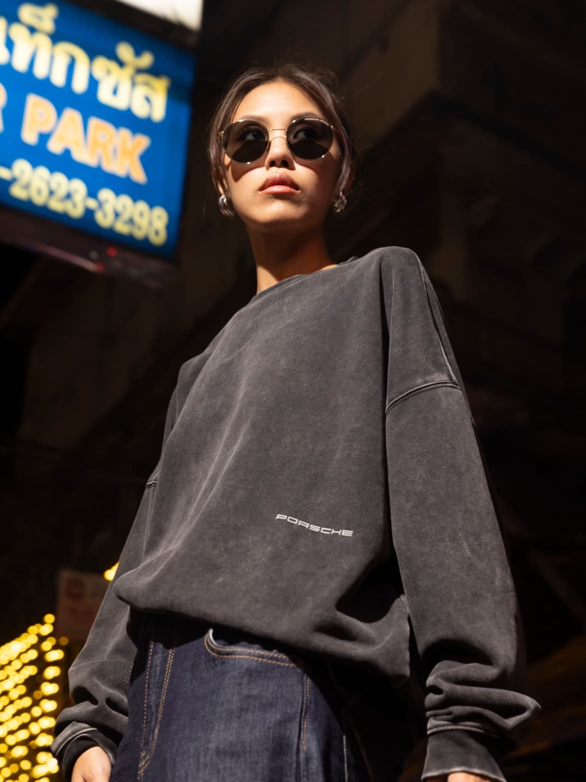 Person in dark Porsche sweatshirt and sunglasses stands confidently in urban night scene with neon lights.