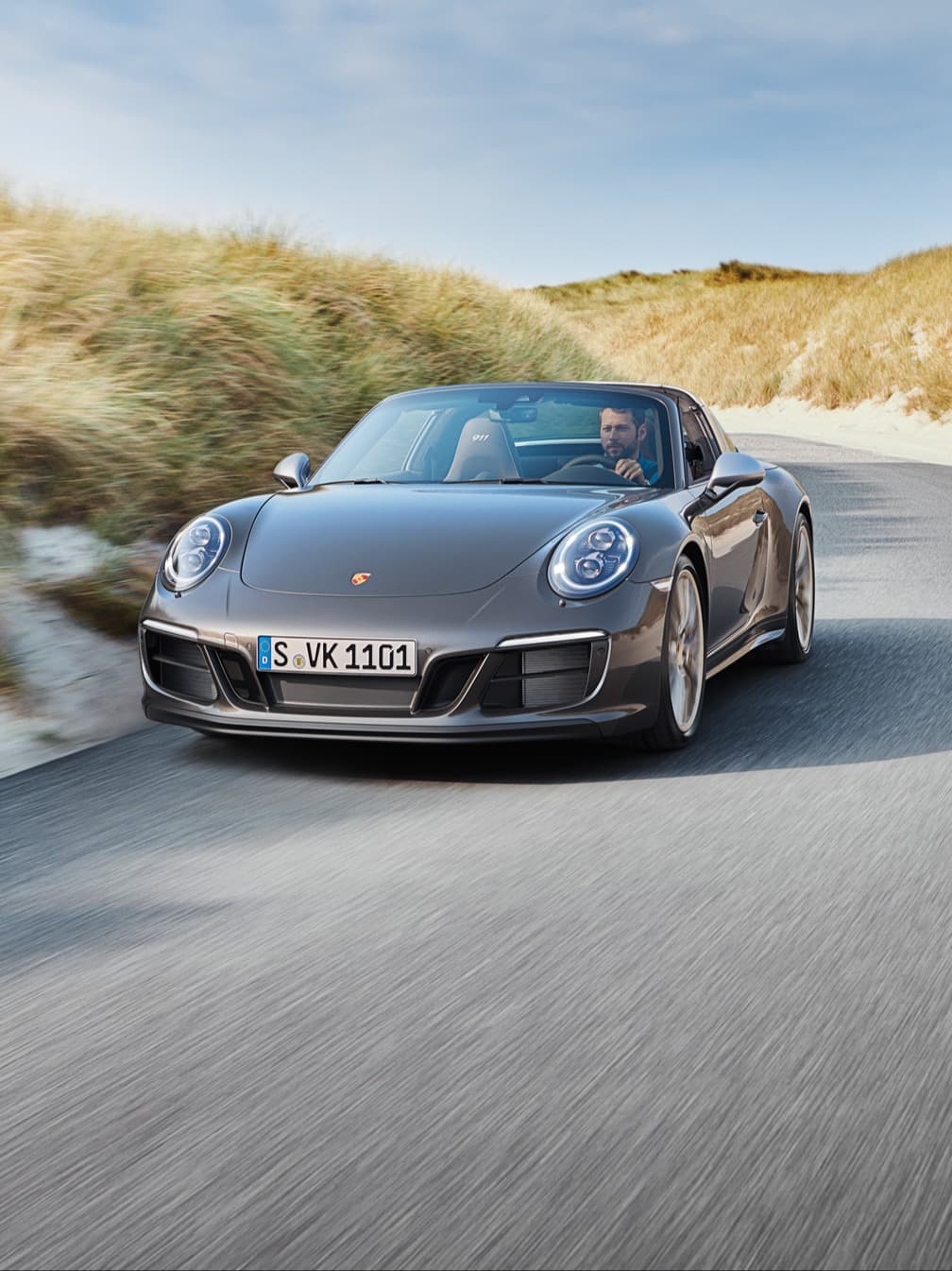 Grijze Porsche 911 Cabriolet rijdt over een kustweg door de duinen, bestuurder achter het stuur, blauwe lucht.