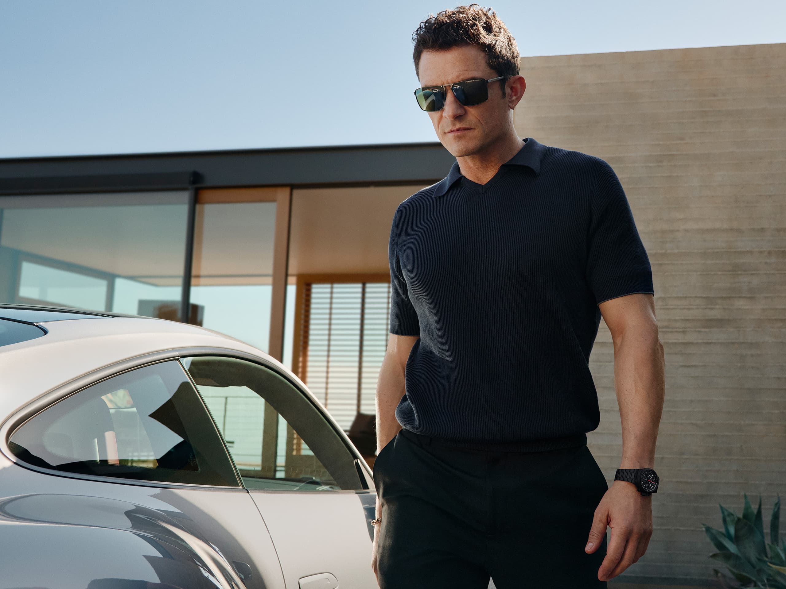 Orlando Bloom wearing the Chronograph 1 - All Black Numbered Edition on his wrist, walking in a carport, his Porsche next to him