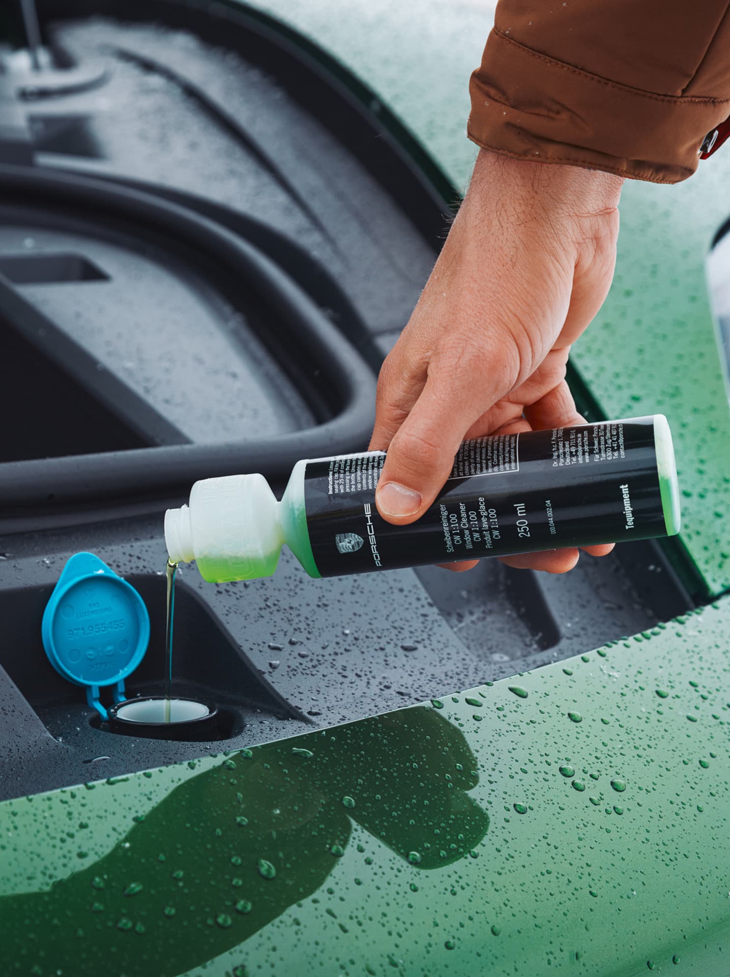 Hand gießt grüne Flüssigkeit aus einer Porsche-Flasche in den offenen Behälter eines Autos, mit Regentropfen auf der grünen Autolackierung.