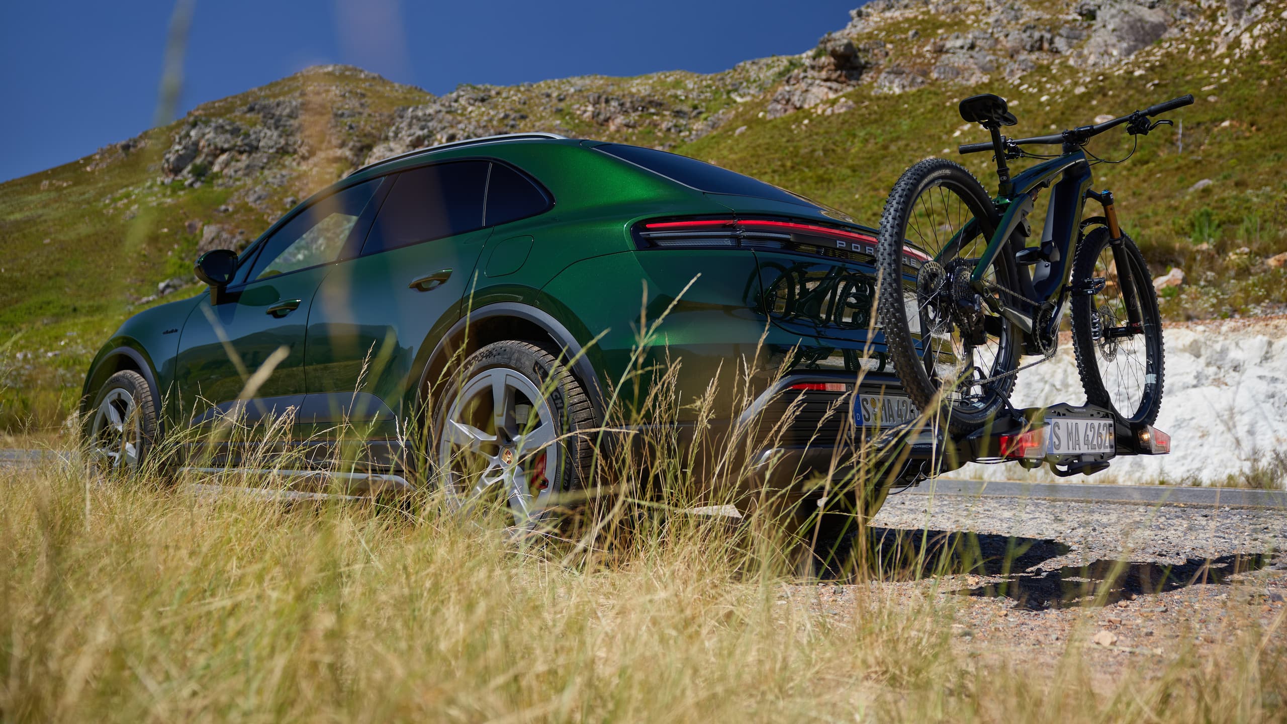 SUV Porsche vert avec porte-vélos sur une route de montagne, entouré d'herbe et de collines rocheuses sous un ciel bleu clair.