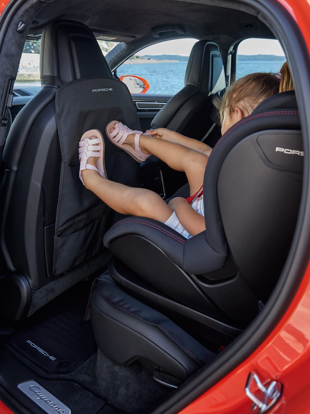 Child in car seat kicks back of front seat in red Porsche, lake view outside.