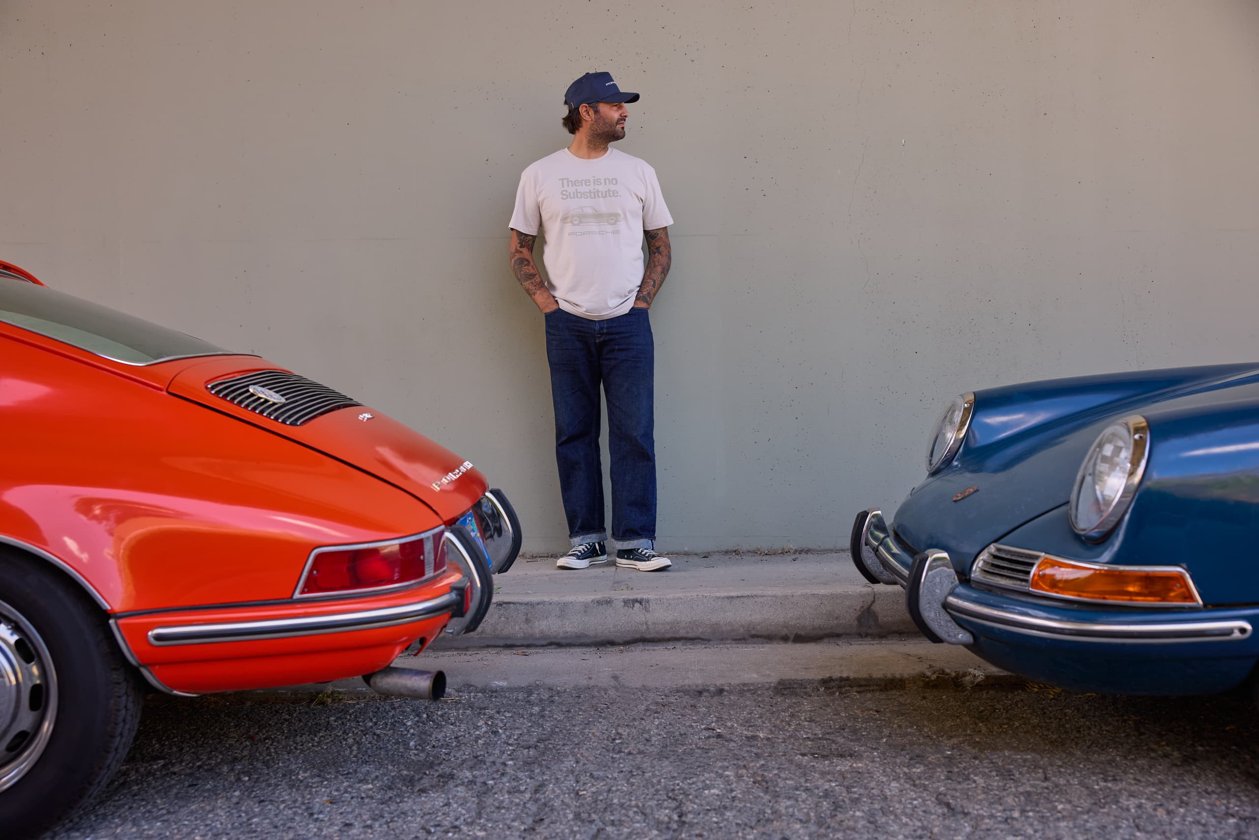 Man in a white Porsche T-shirt and cap standing between a red and a blue Porsche 911 in front of a gray wall.