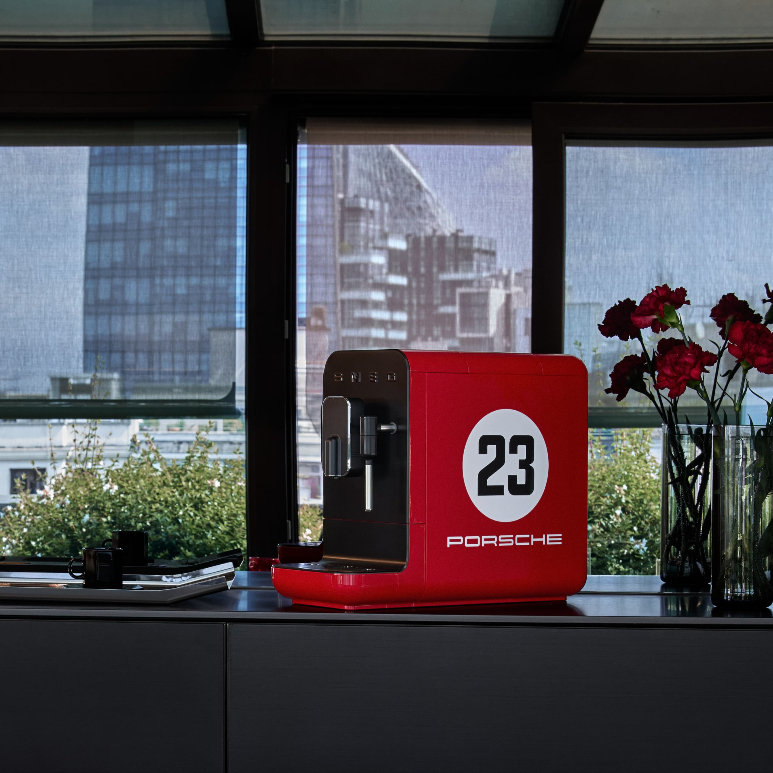 Machine à café Porsche rouge avec le numéro 23, sur un comptoir à côté de fleurs rouges, avec une vue urbaine en arrière-plan.