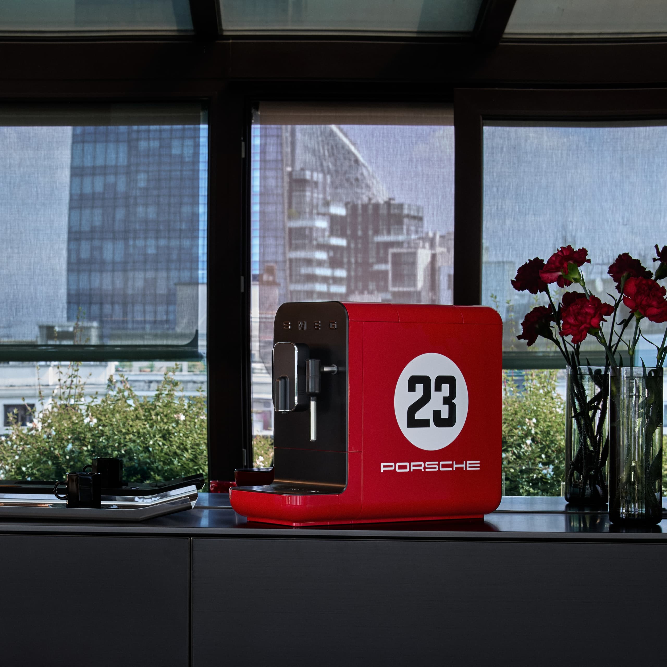 Rode Porsche koffiemachine met nummer 23, op een aanrecht naast rode bloemen, tegen een stadsachtergrond.