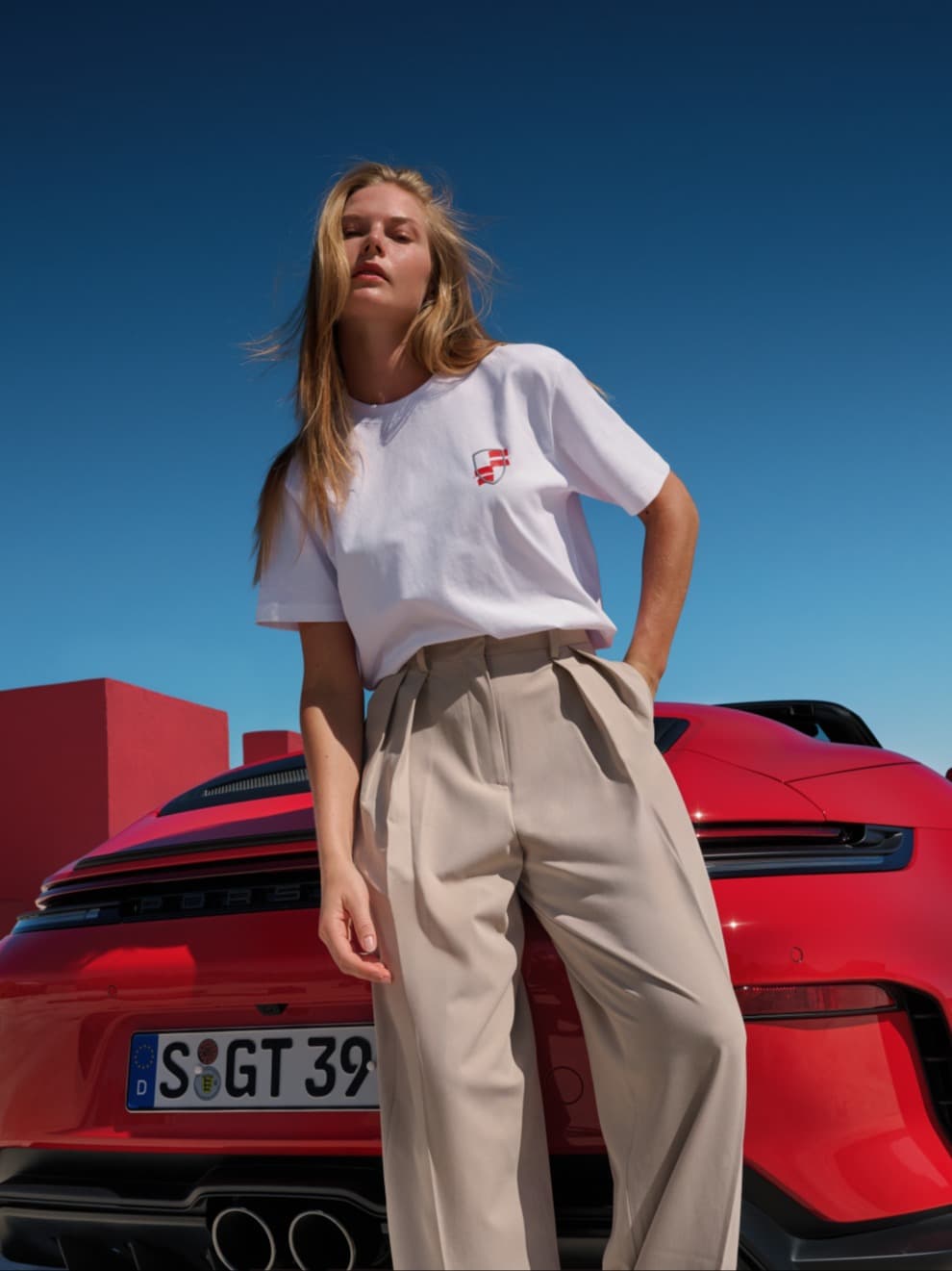 Femme portant un T-shirt blanc et un pantalon beige devant un cabriolet Porsche rouge, ciel bleu en arrière-plan.