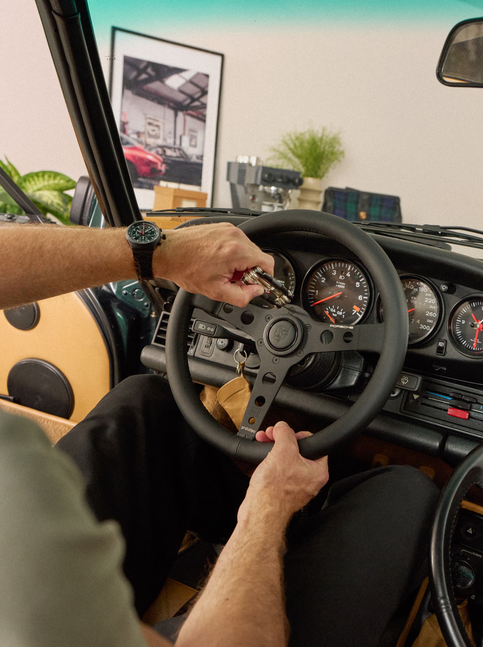 Main tenant portant une montre-bracelet noire, tenant des clés de voiture devant un volant Porsche, avec les compteurs classiques du tableau de bord visibles.