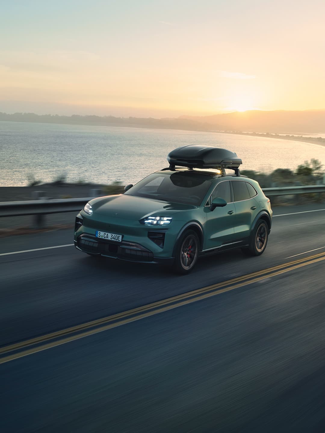 Zöld Porsche SUV fekete tetőboxszal, naplementében egy tengerparti úton haladva.