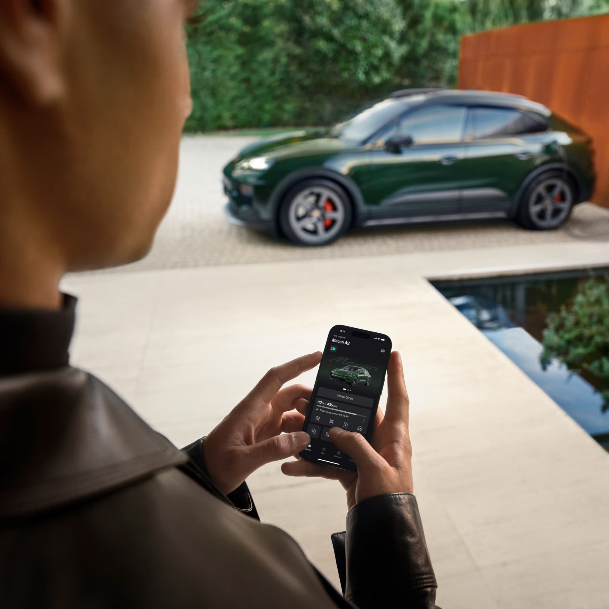 Personne utilisant une application smartphone pour contrôler une Porsche Macan verte garée à l'extérieur, avec une architecture moderne et de la verdure en arrière-plan.