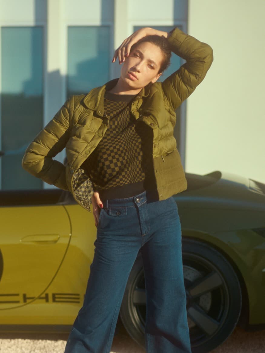 Woman in red Martini Racing jacket and sunglasses sitting by a white car holding collar. Denim jeans and Porsche tumbler v