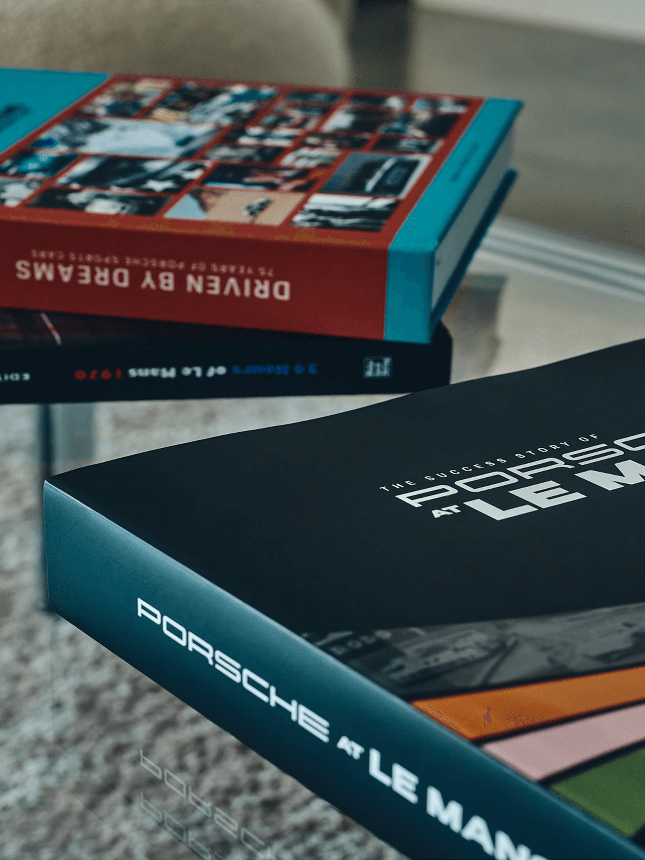 Stapels Porsche-fotoboeken op een tafel, bovenop het rode boek “Driven by Dreams” en het zwarte boek “Porsche at Le Mans”.