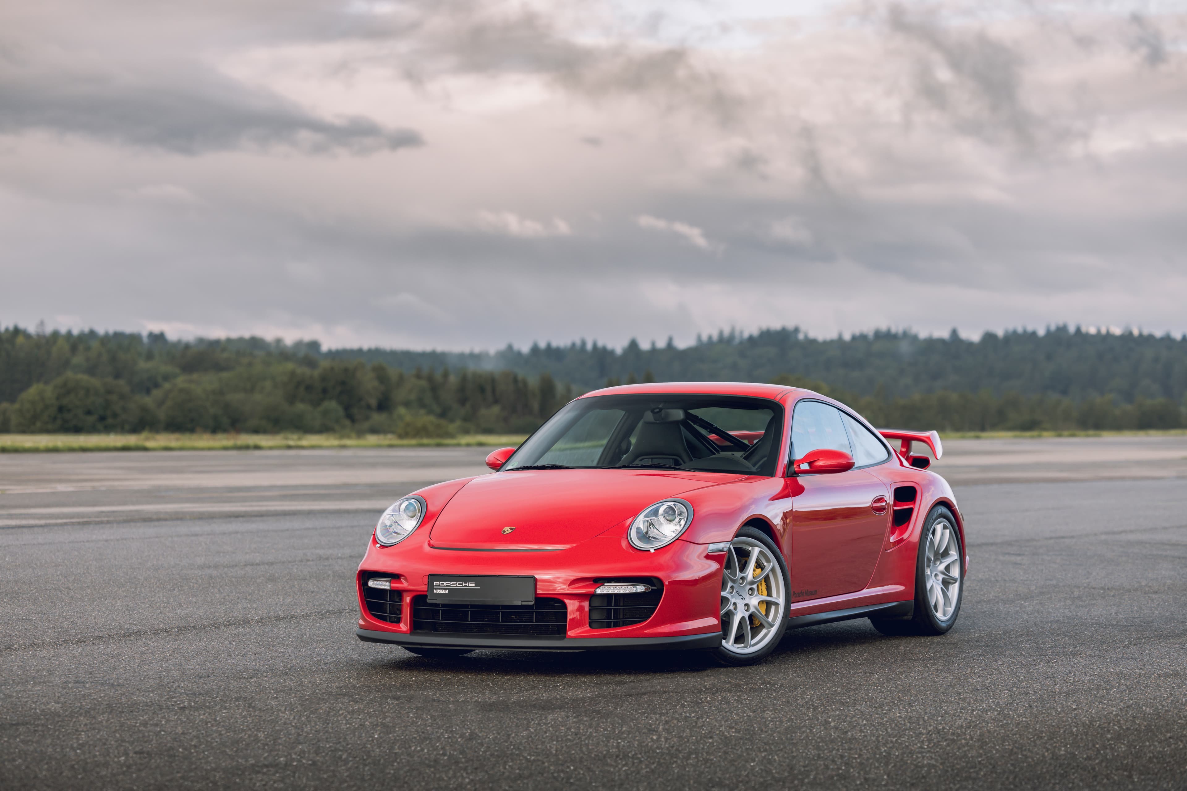 Rode Porsche 911 sportwagen op asfalt met grote achterspoiler, tegen een bosrijke achtergrond onder een bewolkte lucht.