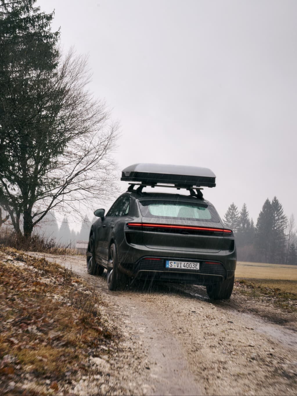 Sort Porsche SUV med tagboks på våd grusvej i regnvejr, træer og grå himmel i baggrunden.