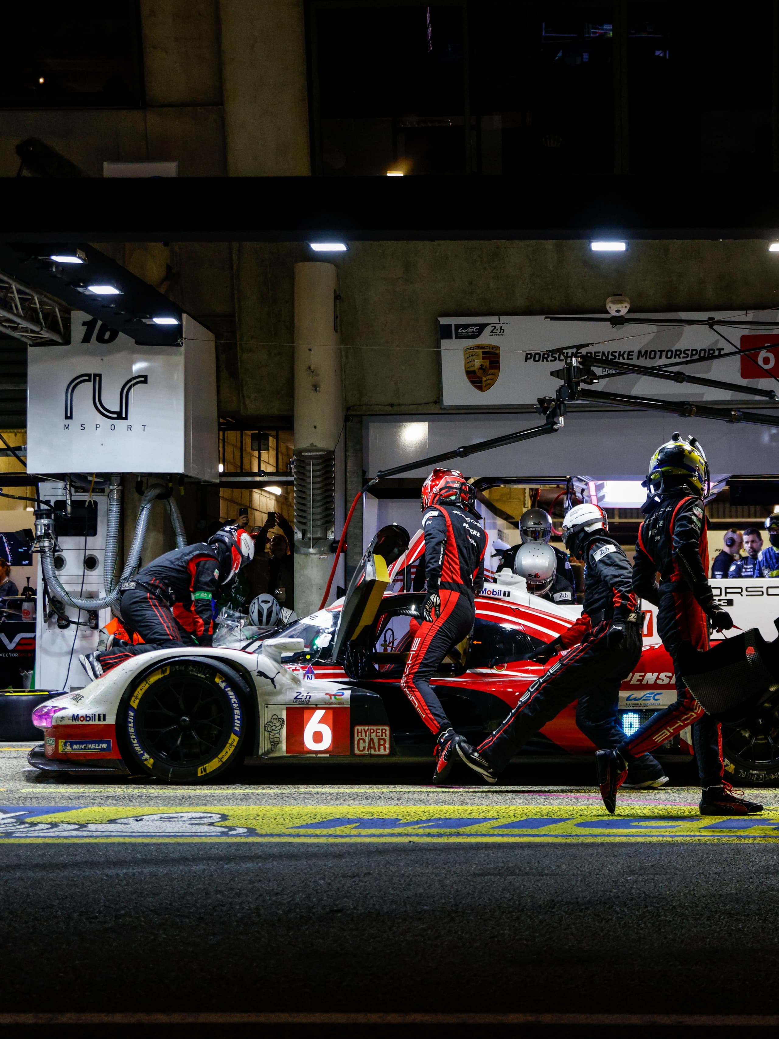 Pitcrew i sorte og røde dragter servicerer en rød-hvid Porsche-racerbil under et natligt pitstop