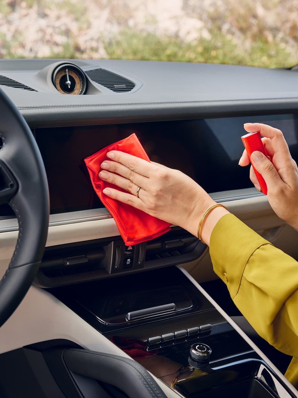 Hand cleaning a Porsche dashboard with a red microfiber cloth and red spray bottle inside the car.