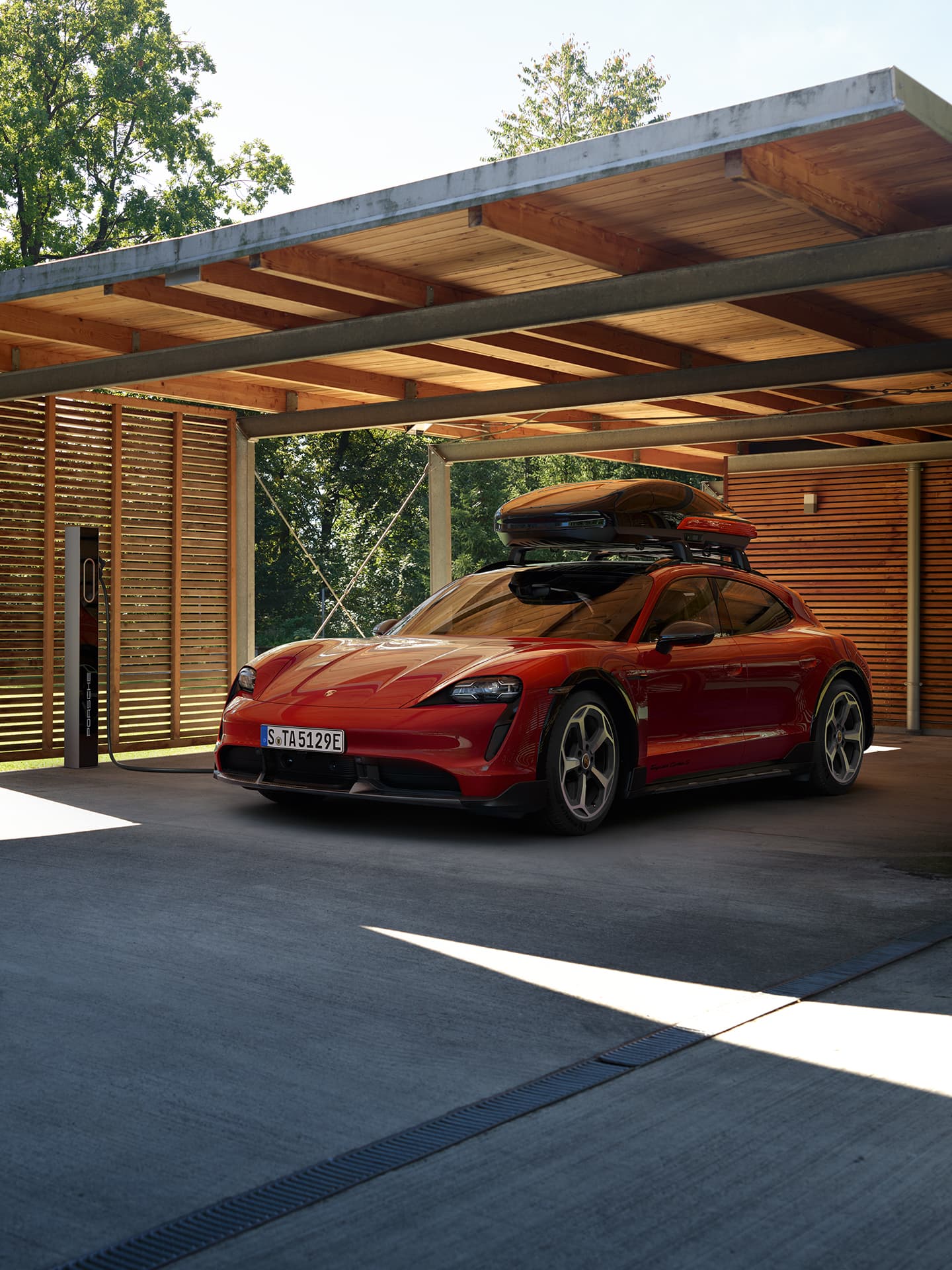 Piros Porsche Taycan Sport Turismo tetőcsomagtartóval, modern fa- és beton autóbeálló alatt, napfényben.