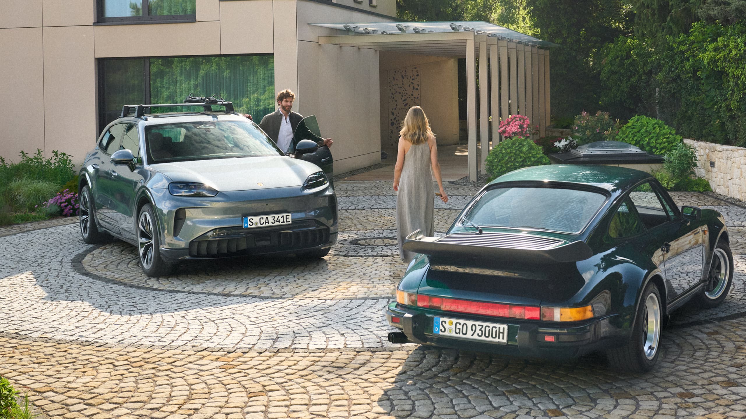 Dva vozy Porsche před moderním domem: šedé SUV se střešním nosičem a zelený model 911; muž vystupuje, žena jde ke dveřím.