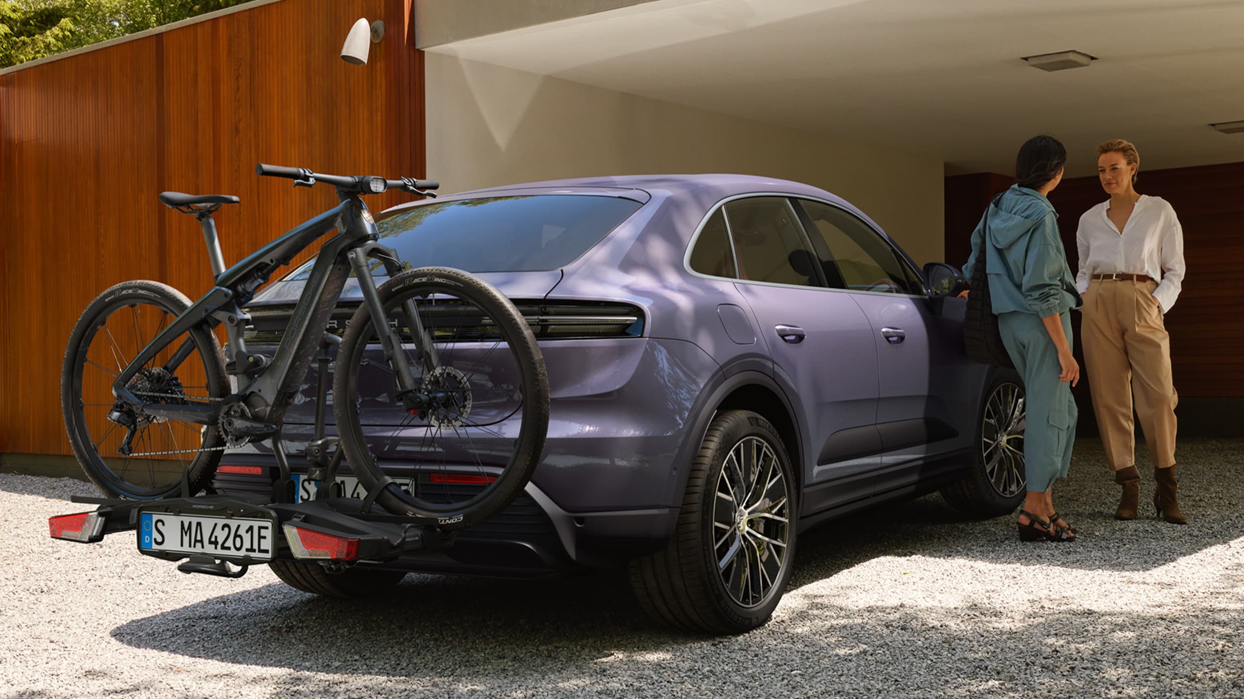 Dwie kobiety rozmawiają, stojąc obok elektrycznego Macana w kolorze prowansalskim; z tyłu na bagażniku rowerowym zamocowany jest rower Porsche eBike.