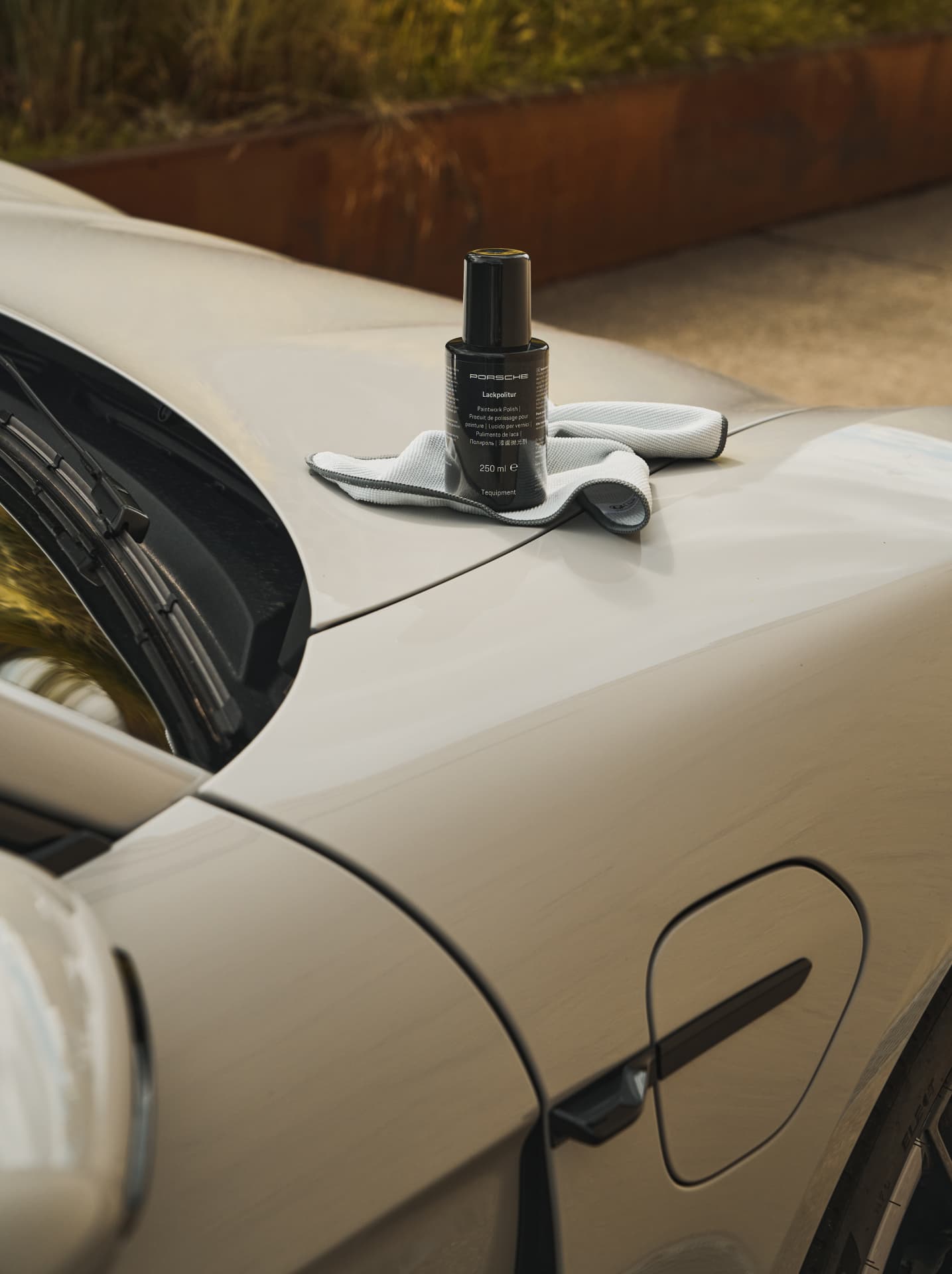 Porsche-Politurflasche auf einer grauen Motorhaube mit einem Mikrofasertuch, im Freien mit verschwommener grüner Kulisse.