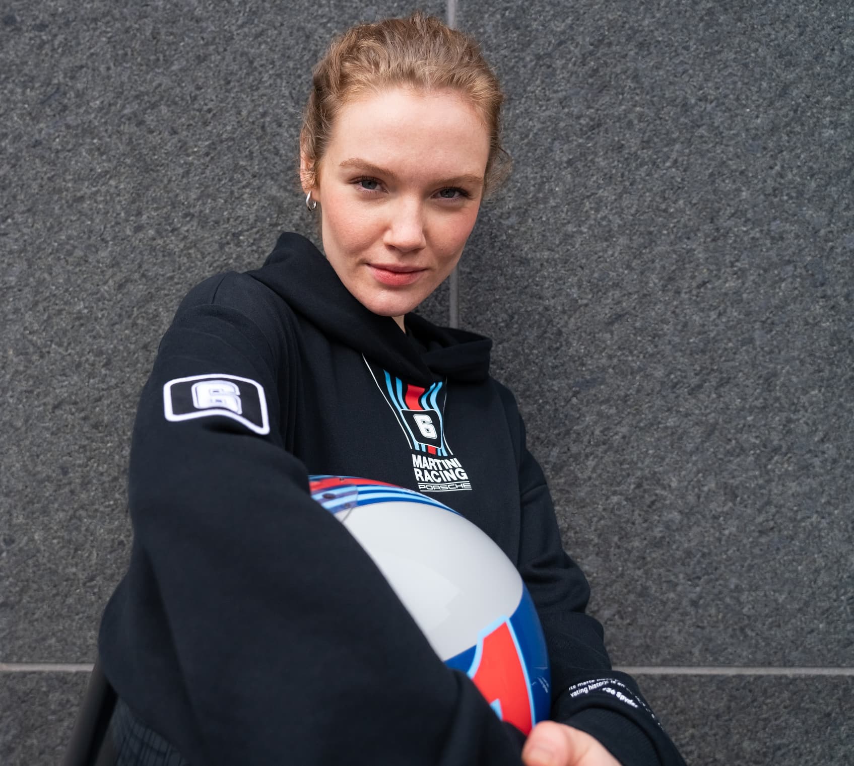 Woman leaning against a wall wearing hoodie from the Porsche x MARTINI RACING collection holding a helmet under her arm.