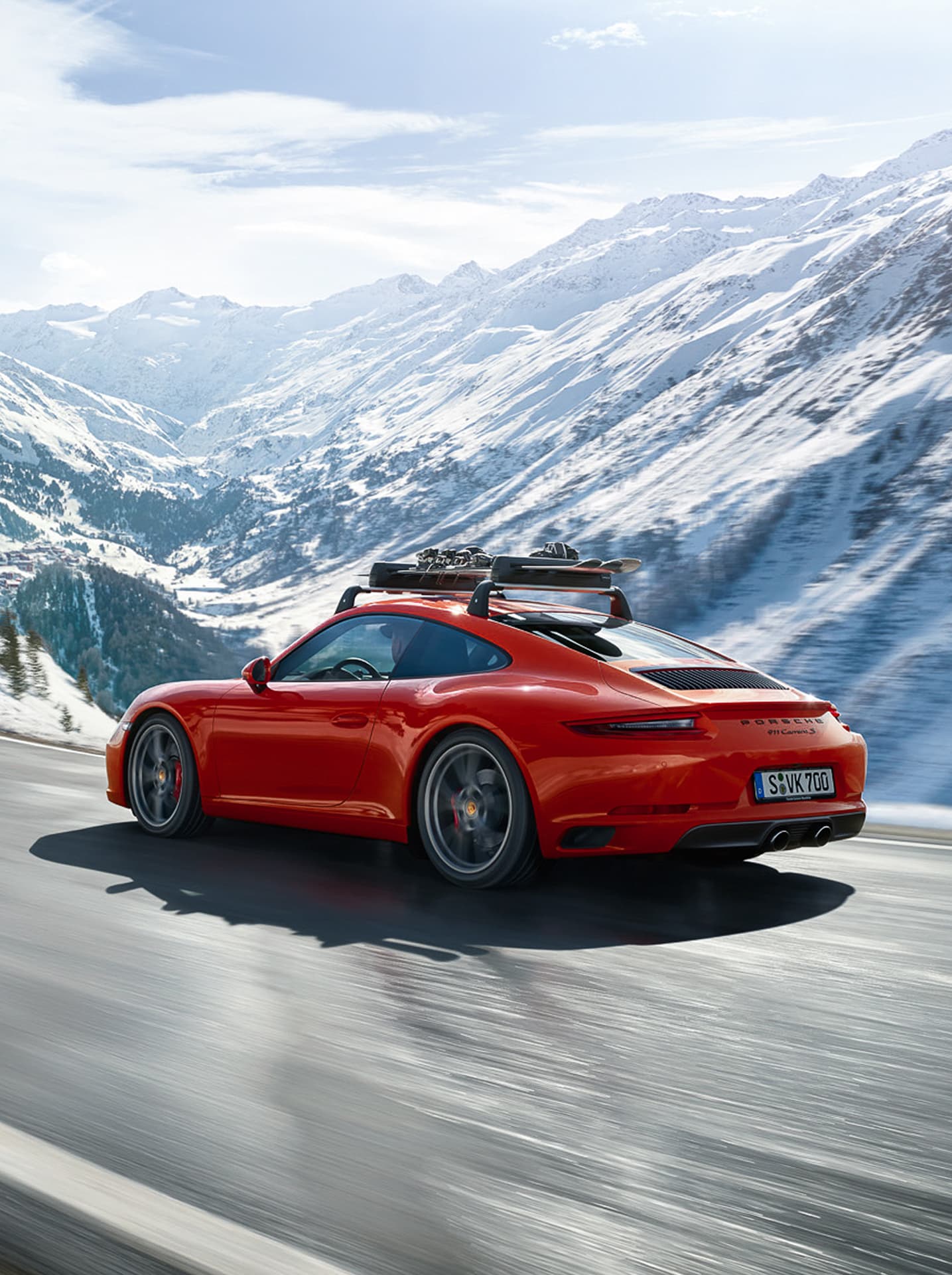 Roter Porsche 911 Carrera mit Dachgepäckträger auf verschneiter Bergstraße unter klarem Himmel.