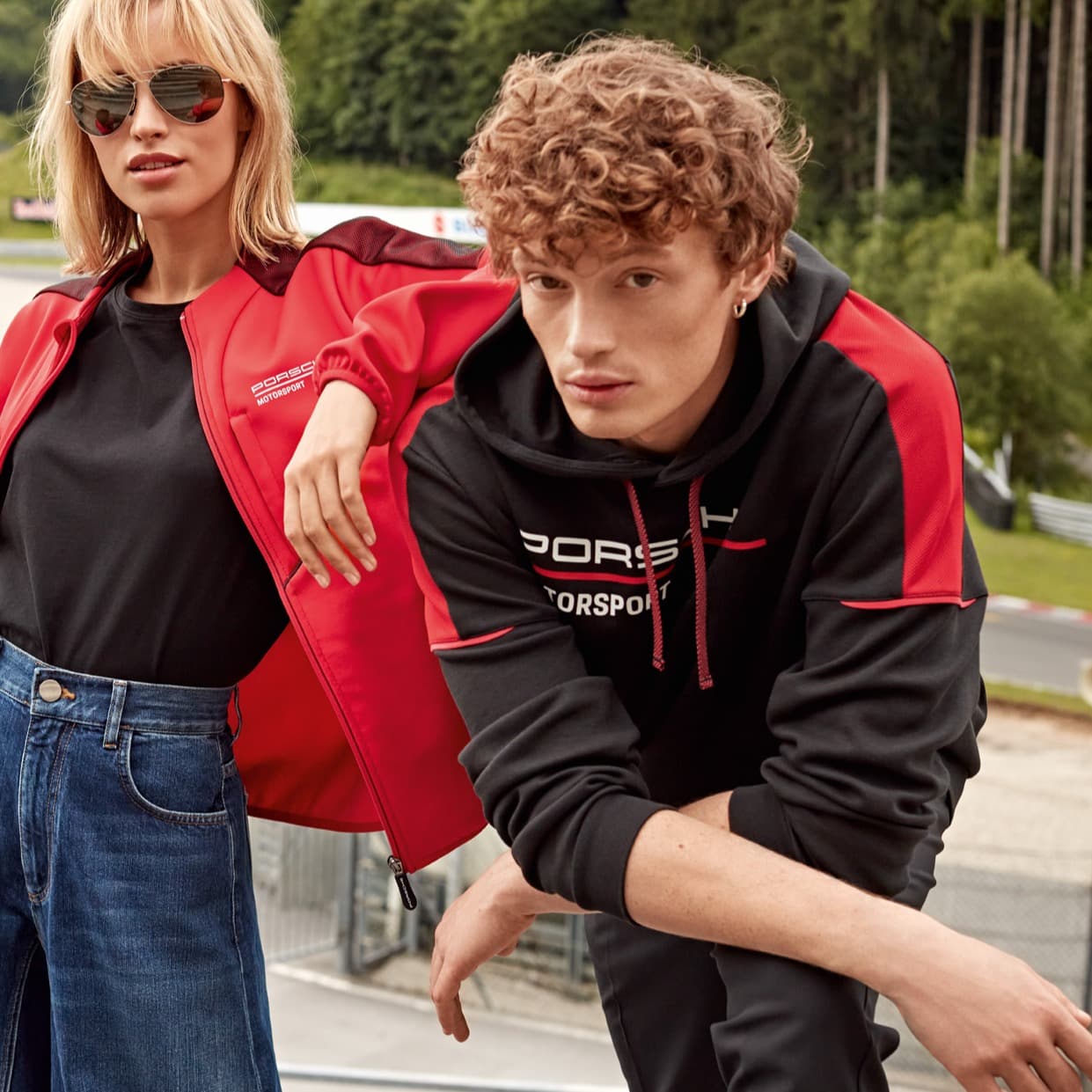 Zwei Modelle in Porsche Motorsport-Bekleidung, rote Jacke und schwarzer Hoodie, im Freien mit einer Rennstrecke im Hintergrund.