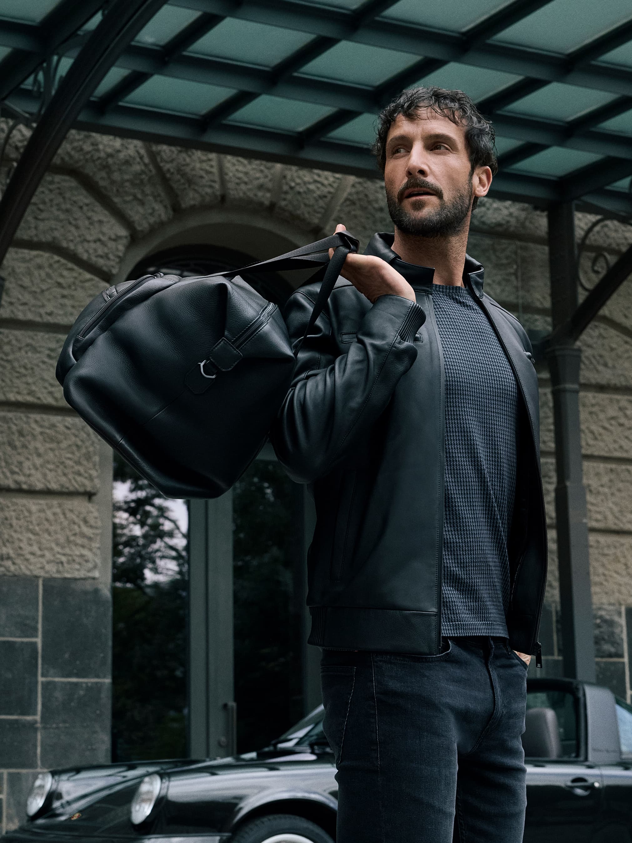 Man in black leather jacket holding a black duffel bag, standing in front of a classic car under a glass canopy.