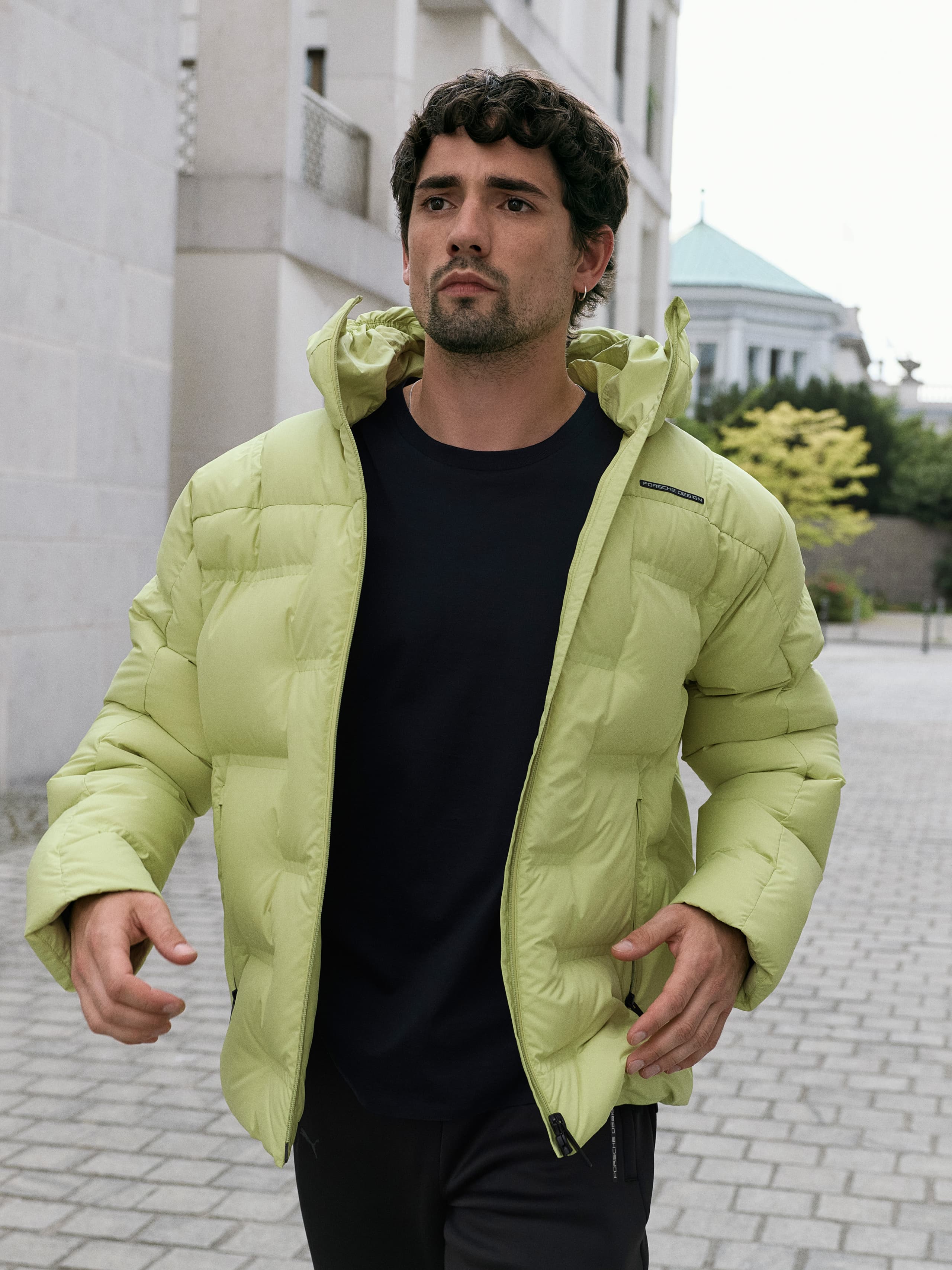 Man in a light green puffer jacket walking on a city street, wearing a black shirt, with buildings and trees in the background.