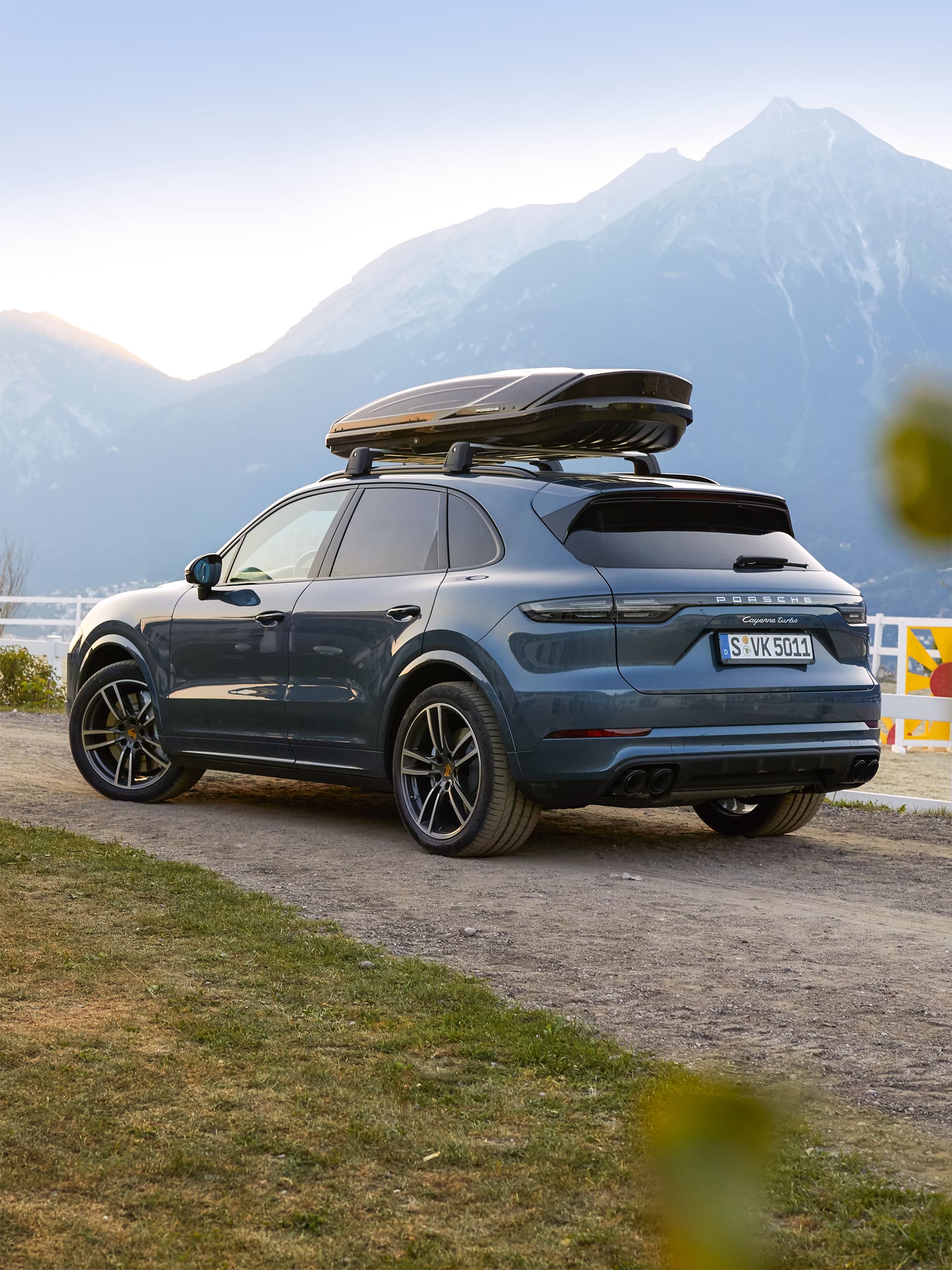 Šedé Porsche Cayenne s černým střešním boxem, zaparkované na štěrkové cestě před horským panoramatem v podvečerním světle.