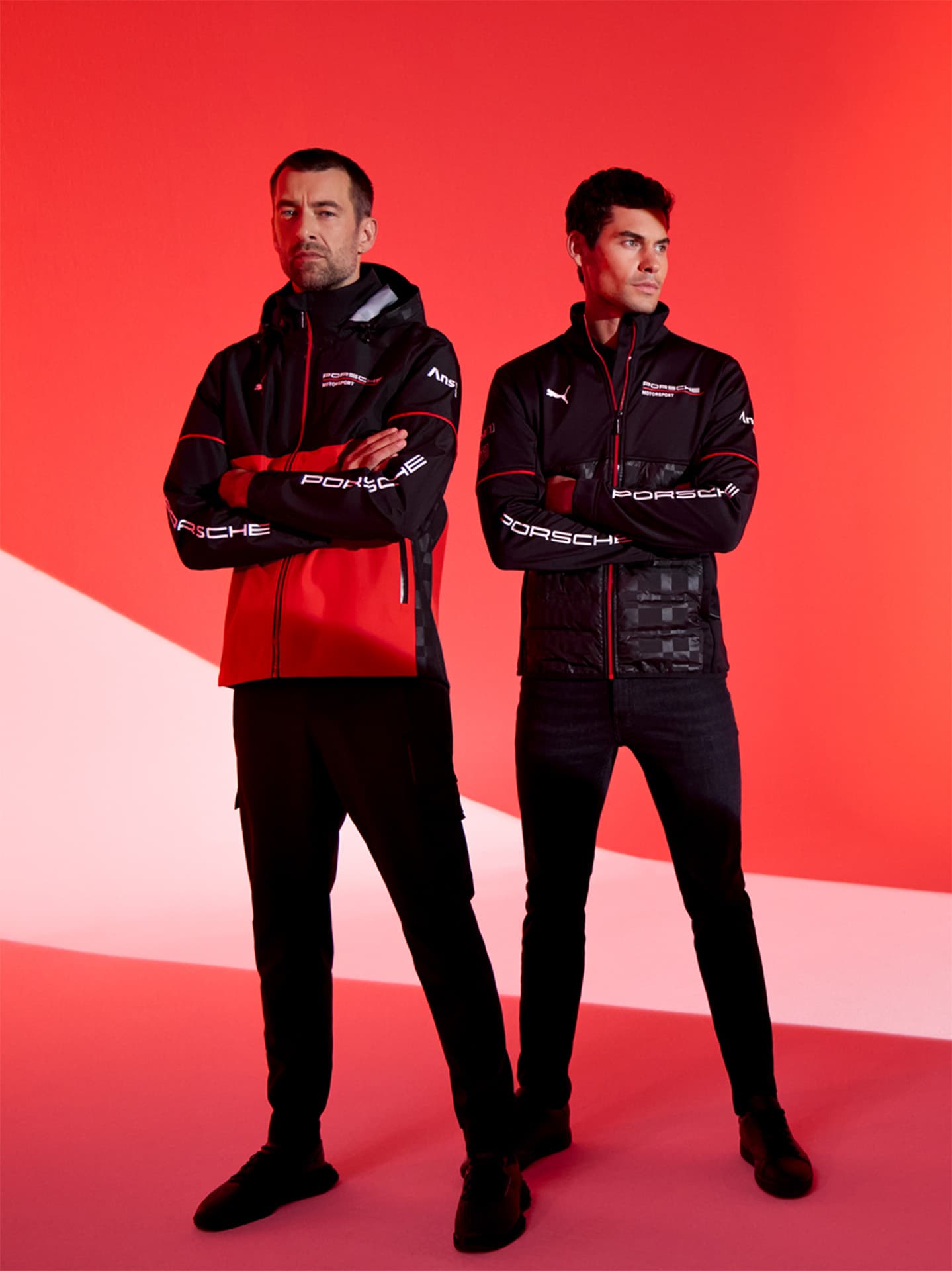 Two people wearing Porsche Motorsport clothing and sunglasses at the racetrack fence under a blue sky.